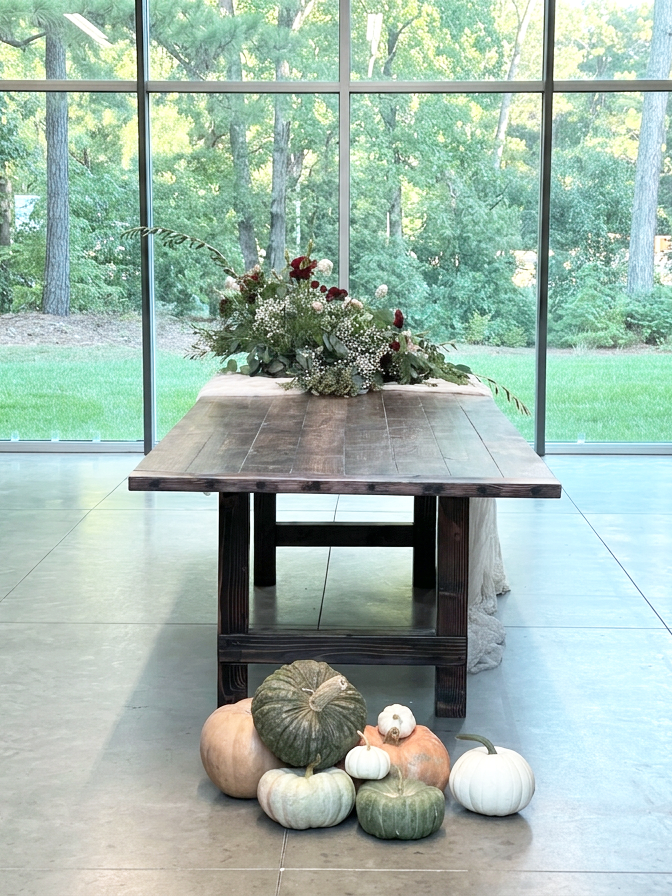 A wood table with a flower arrangement on top, positioned in front of large glass windows showing a green, wooded outdoor area. Several pumpkins and gourds are arranged at the base of the table on the floor.