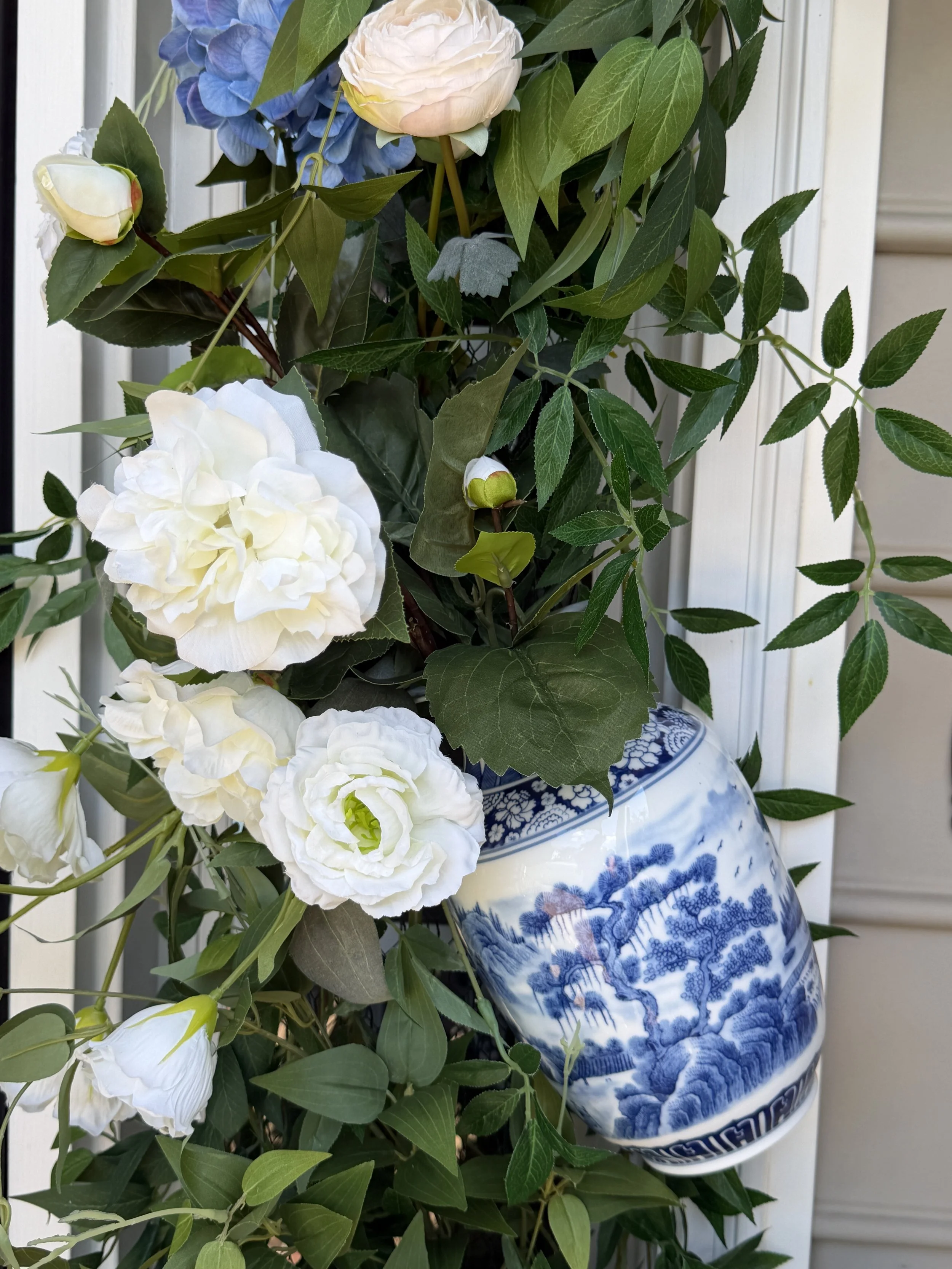 A large decorative blue and white porcelain vase with floral patterns, holding a bouquet of white and light pink flowers accompanied by green leaves, placed outdoors against a white siding wall.