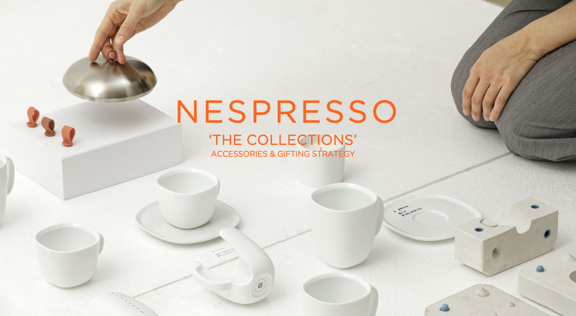 Various white ceramic cups, saucers, and accessories, with a metallic bell-like object being placed on a white block, and a person kneeling nearby, suggesting a collection display for Nespresso accessories.