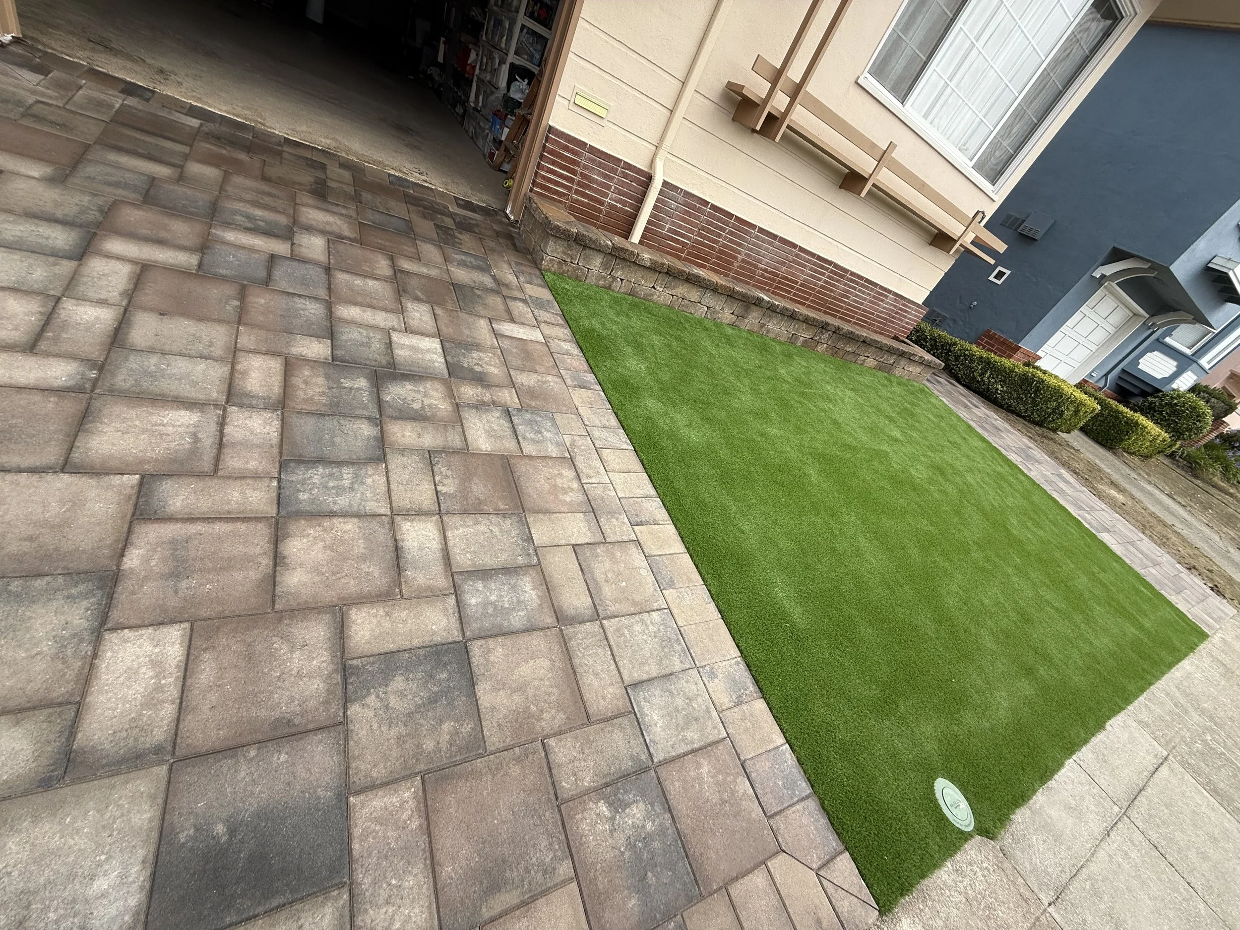 A backyard with a newly installed artificial grass lawn, adjacent to a brick-paved driveway and a beige house with a window, a small garden with trimmed bushes, and a neighboring house with a blue wall.