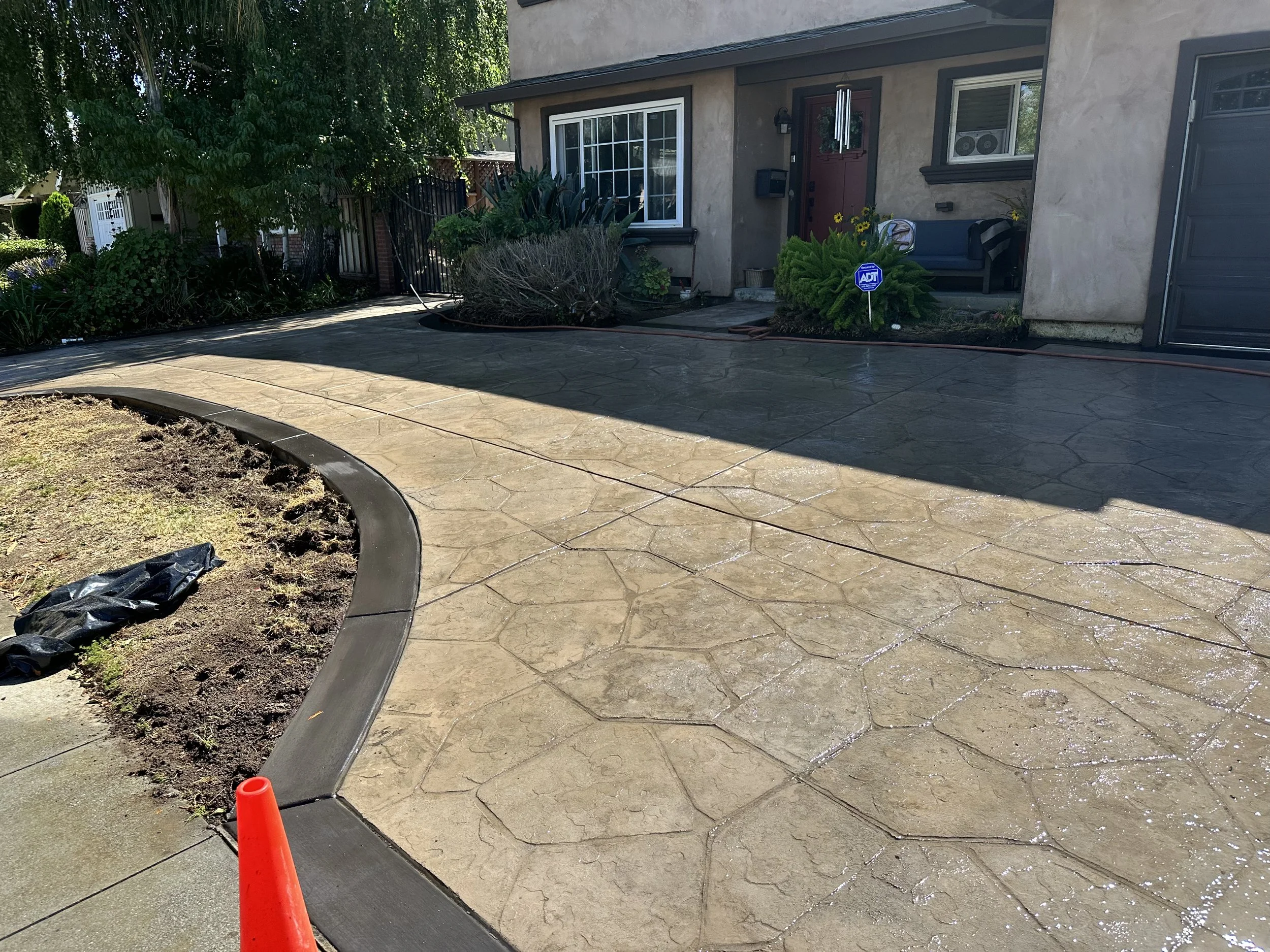 Recently poured concrete driveway with decorative patterns in front of a house, with some garden beds, plants, and a porch with a bench and plants nearby.