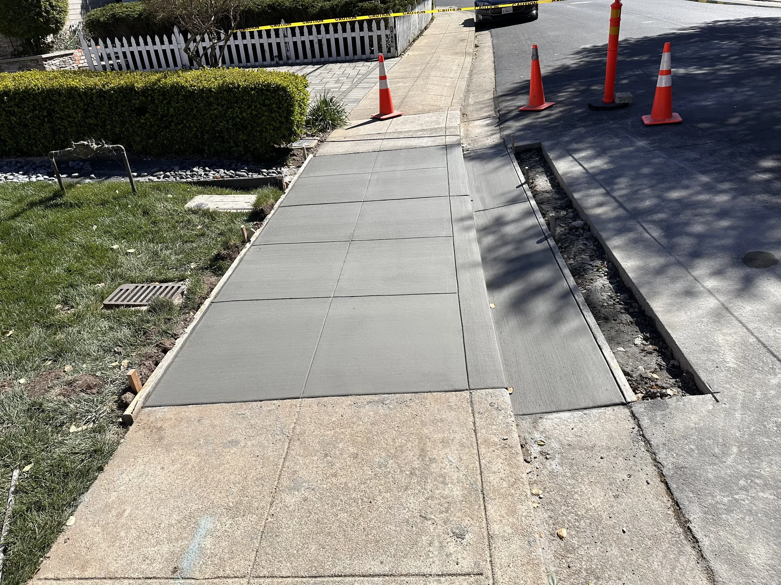 Newly poured concrete sidewalk on a residential street; orange construction cones and caution tape blocking off the part of the road; green grass and bushes on the left side of the sidewalk.