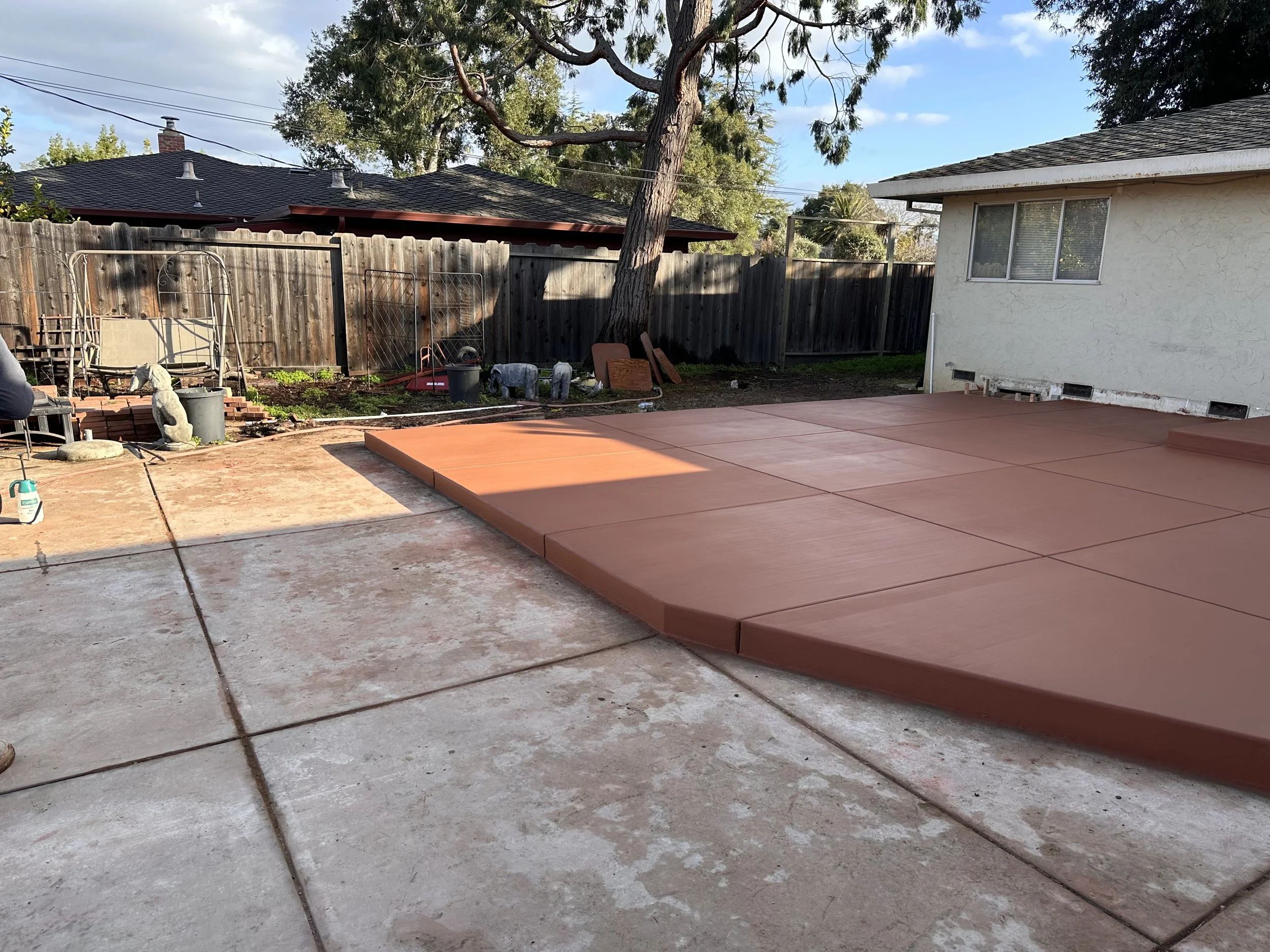 Backyard patio undergoing renovation with new poured concrete and a section covered in reddish-brown material, in front of a white house with a window, surrounded by a wooden fence, trees, and outdoor furniture.