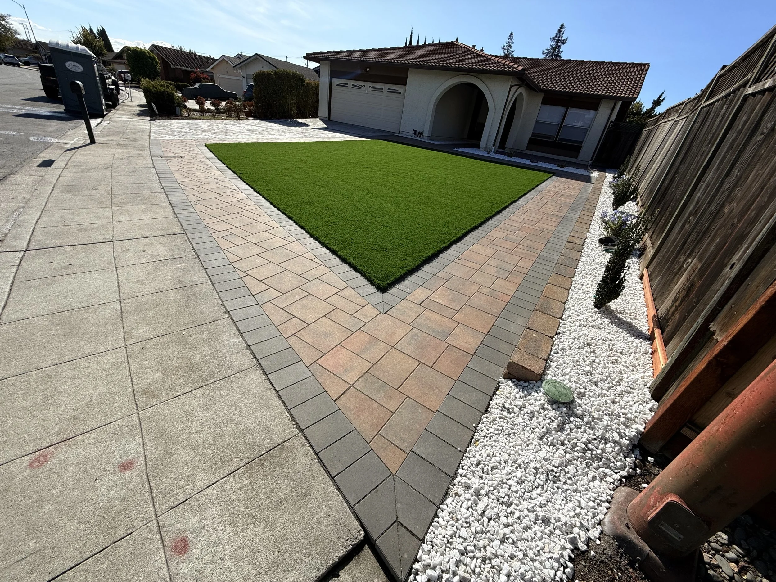 Front yard with green lawn, paved walkway, and flower bed with white rocks and plants next to a wooden fence.
