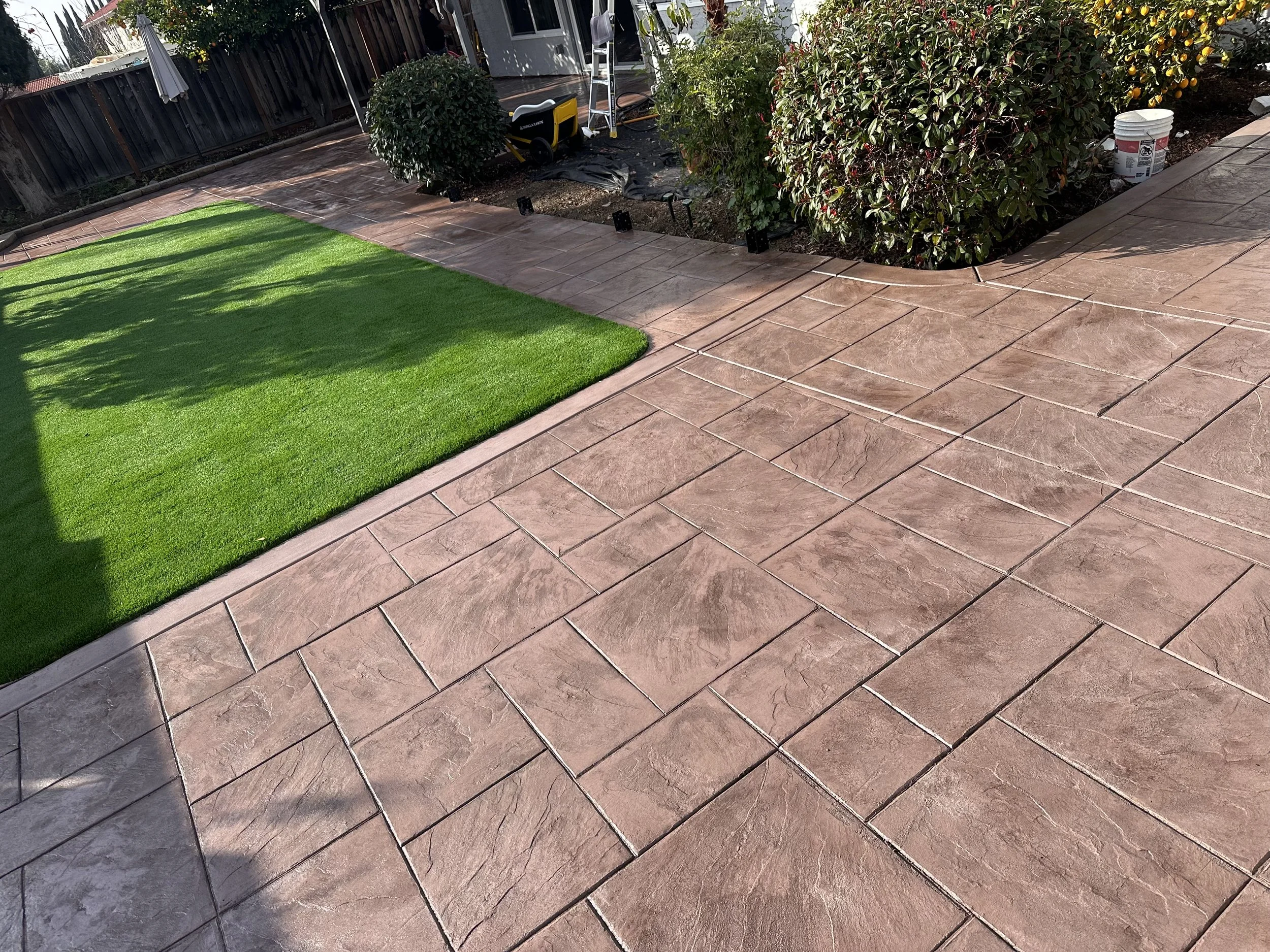 Newly installed pinkish-brown stamped concrete patio with a strip of bright green artificial turf on the left, surrounded by a garden with bushes and a white bucket.