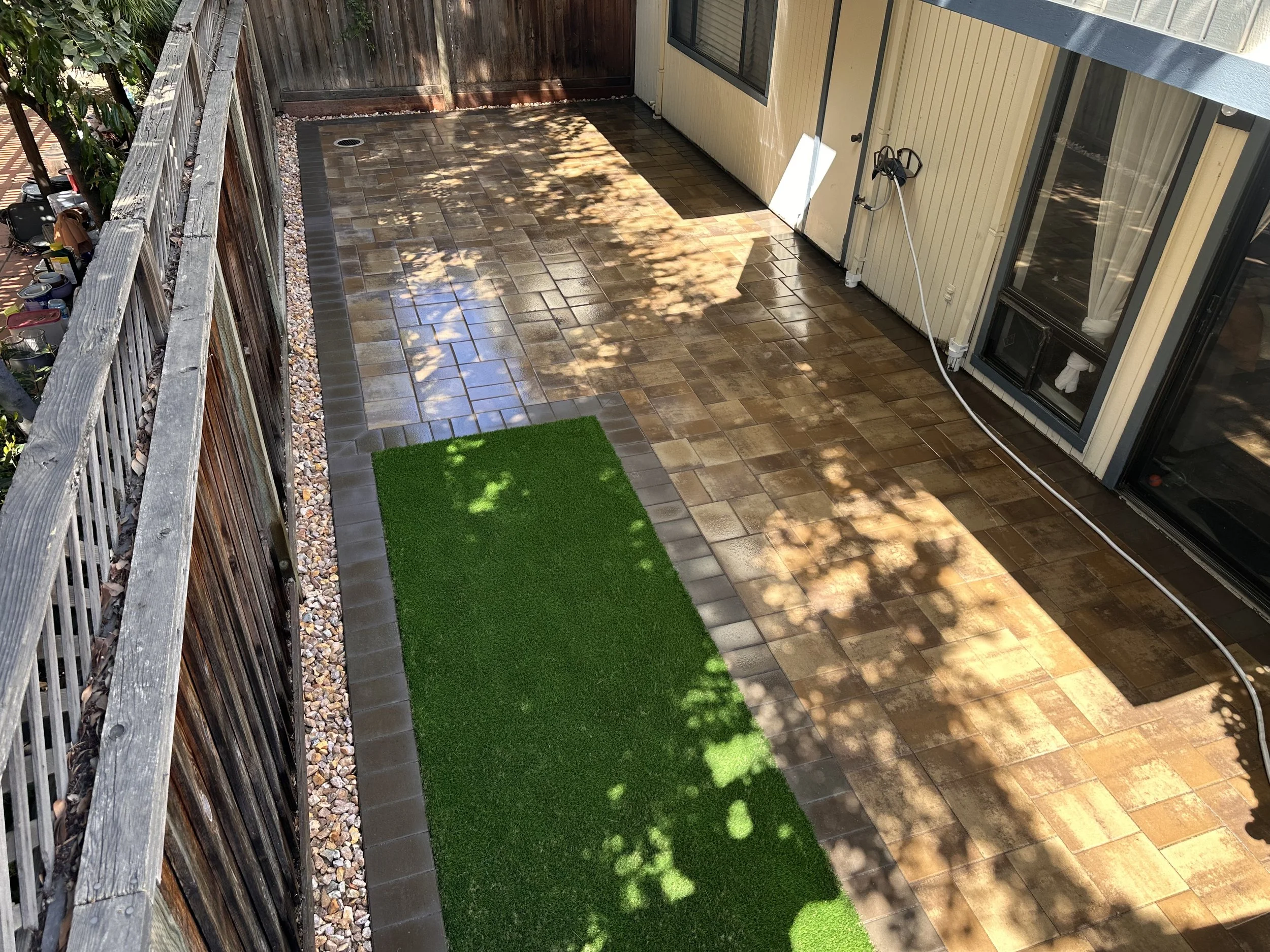 Backyard patio with wet, brown, rectangular tiles, a green artificial grass mat, and a wooden fence with plants, with shadows of trees on the ground.