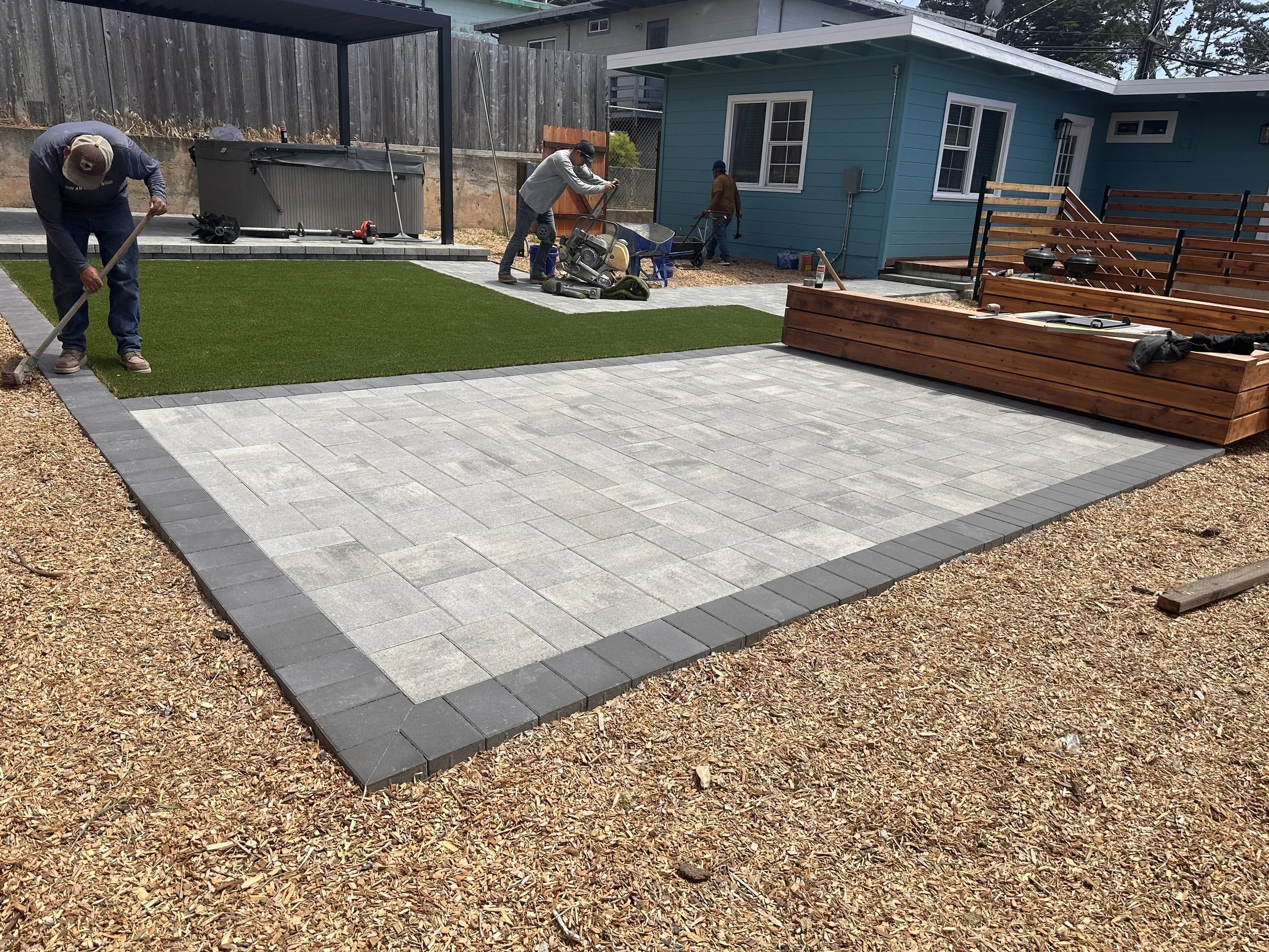 People building a backyard patio with gray pavers, a small patch of artificial grass, and a raised wooden deck. One person is raking the gravel, another is using a power saw, while others work near a blue house.