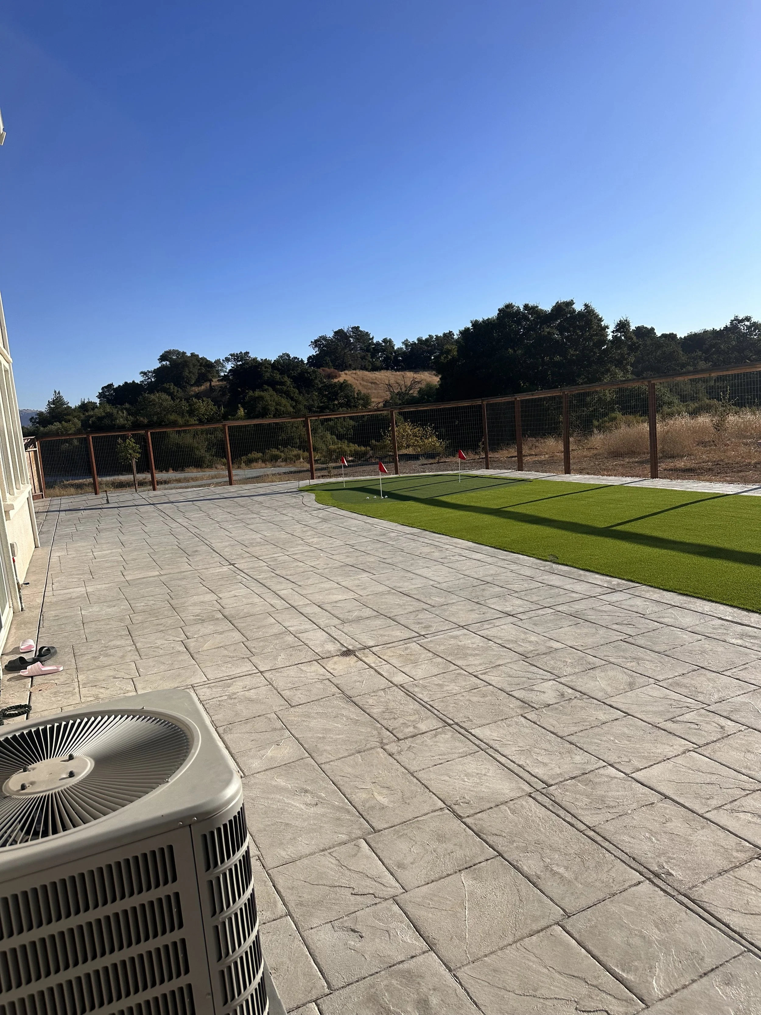 Patio area with paver stones, green artificial turf, and a obstacle golf net with red flags in the background under a clear blue sky.