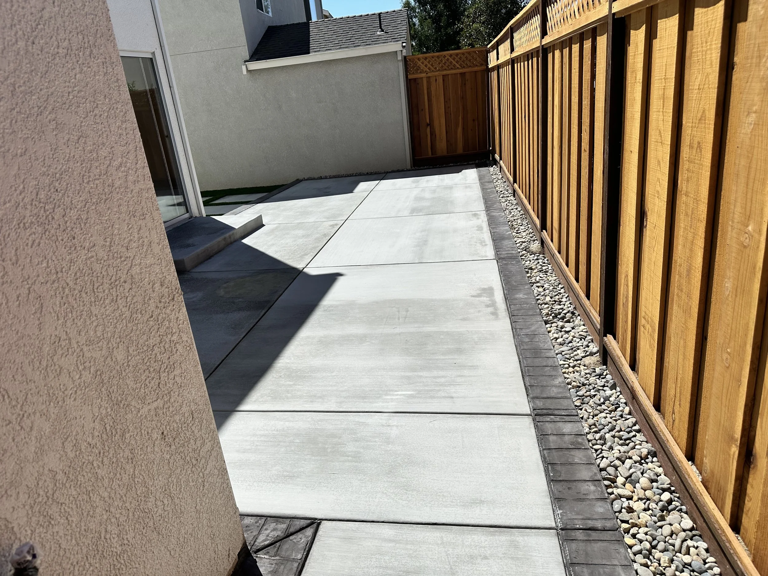 Concrete patio next to a wooden fence and a beige stucco wall of a house with a sliding glass door, in bright sunlight with shadows.
