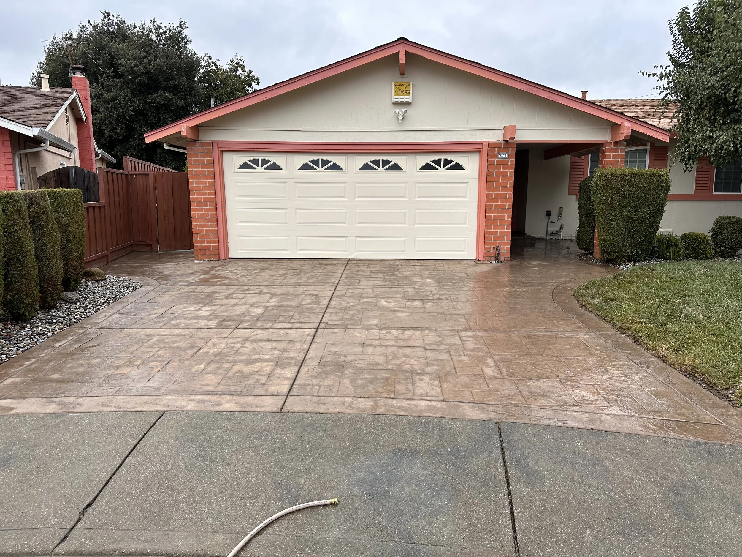 Front view of a house with a white double garage door, red brick accents, and a concrete driveway that appears wet, with a green grassy area and bushes on the right side.
