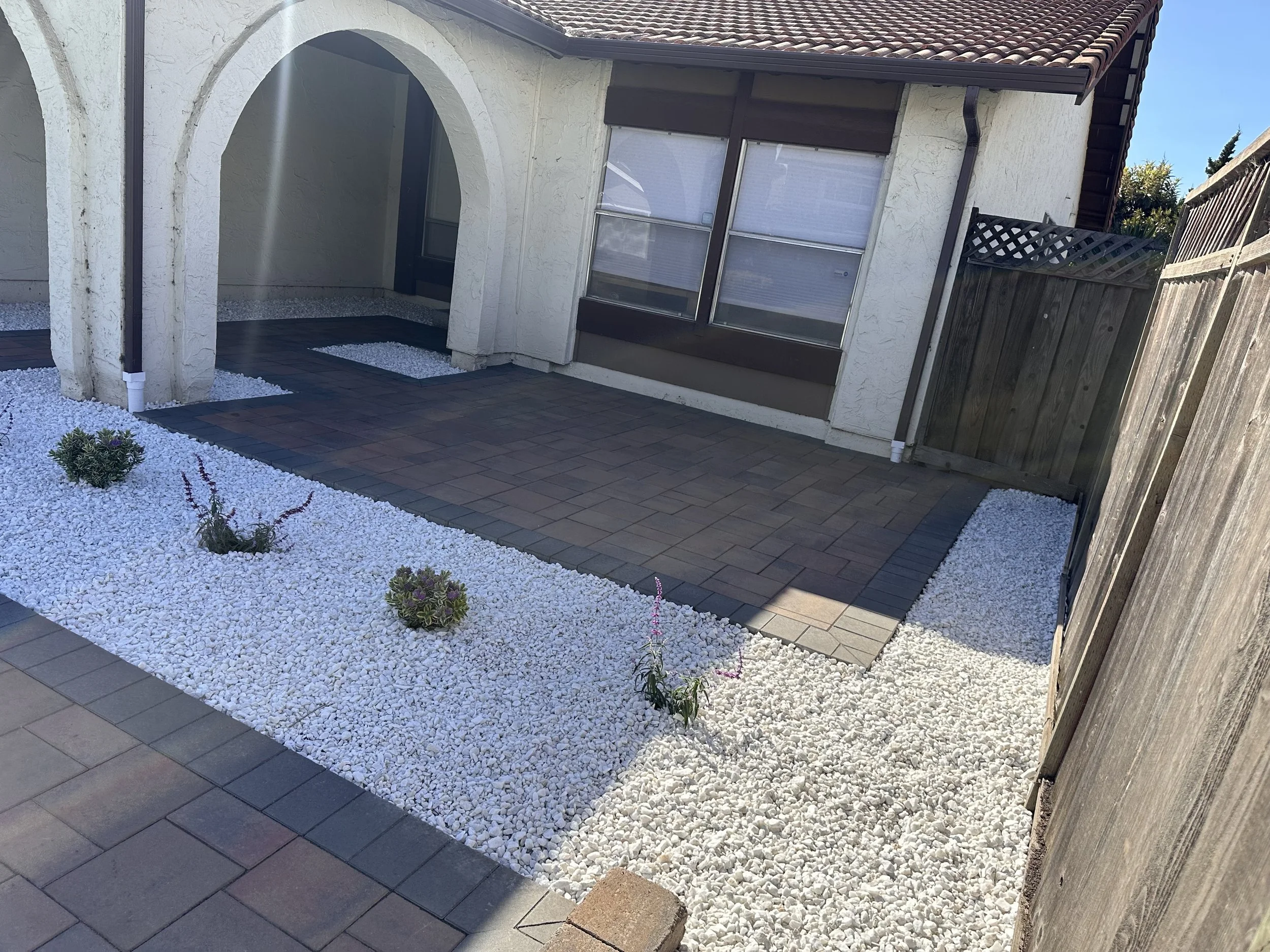 Backyard patio with brown pavers, white gravel beds, small plants, and a white stucco house with windows, an archway, and a wooden fence.