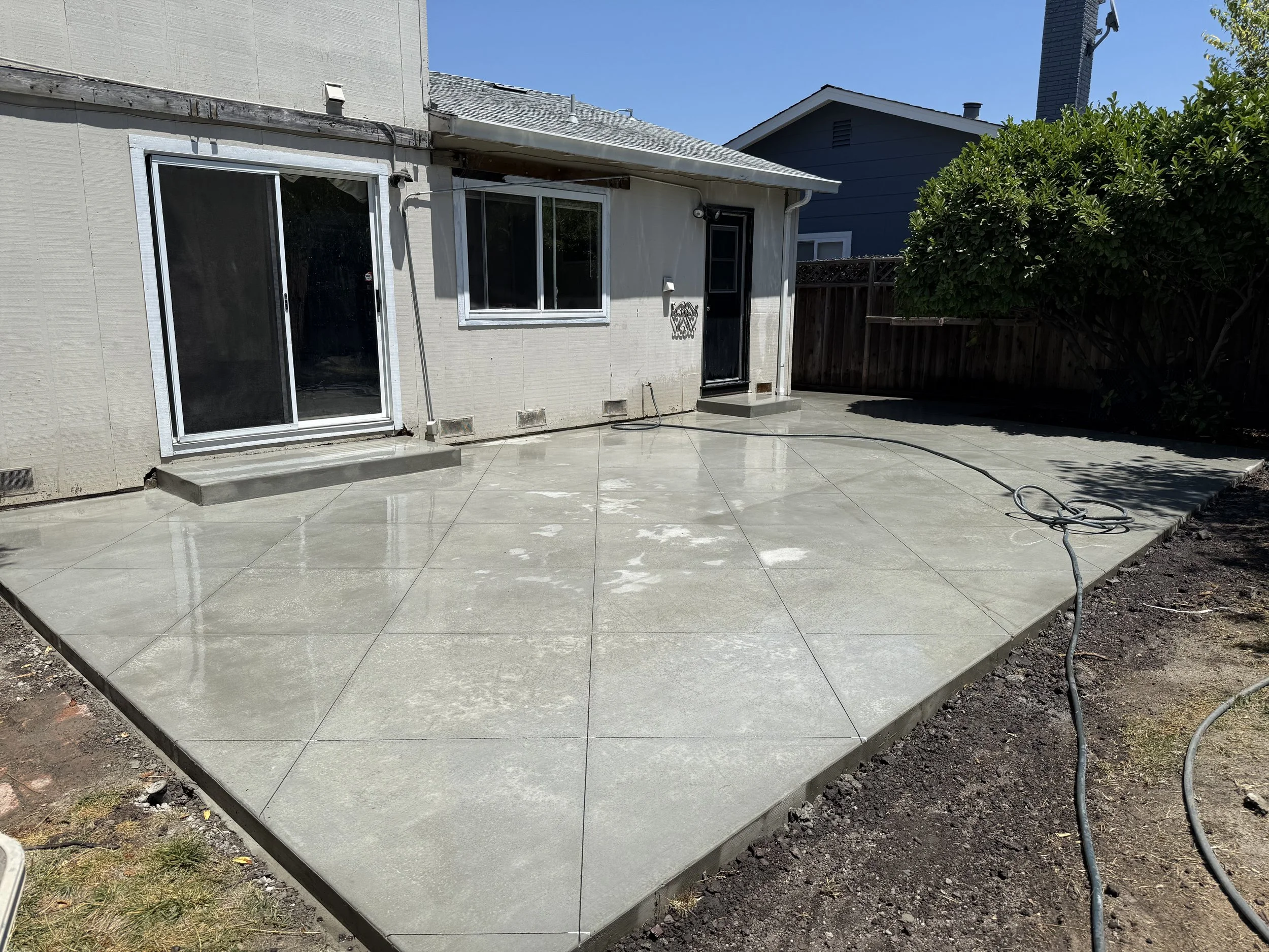 Finished concrete patio with a rolling hose on it in the backyard of a house with sliding glass door and window, fenced yard, blue neighboring house, and surrounding trees.
