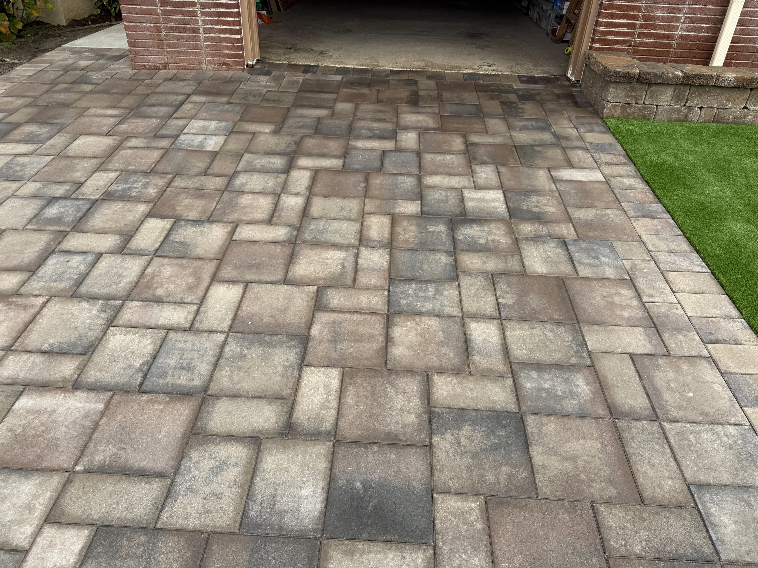 Decorative paver driveway with a parking space adjacent to a brick house, with a small patch of artificial grass and a brick retaining wall.