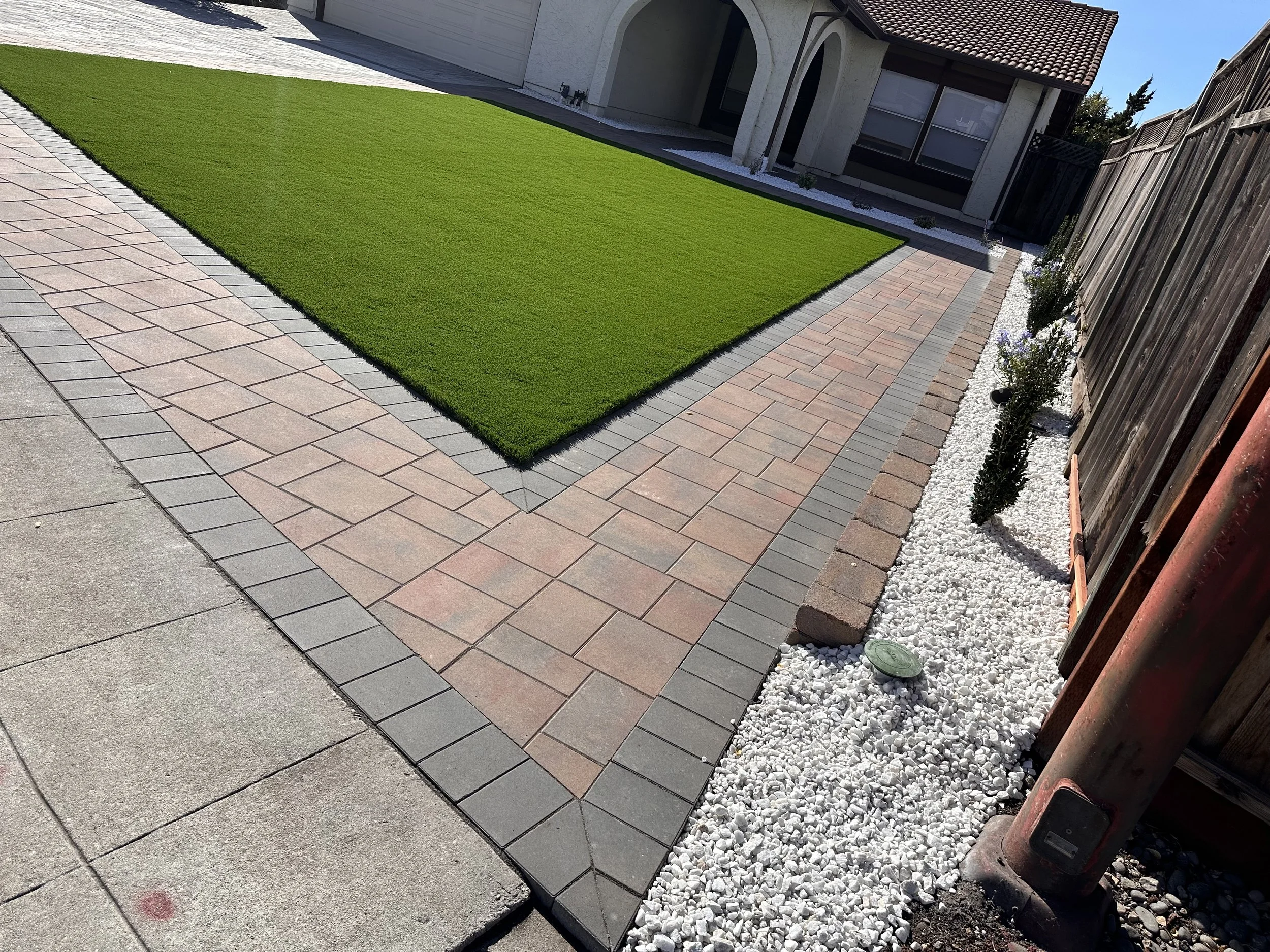 A neatly landscaped backyard with a green grass lawn, brick walkway borders, a gravel strip with small plants, and a house in the background.