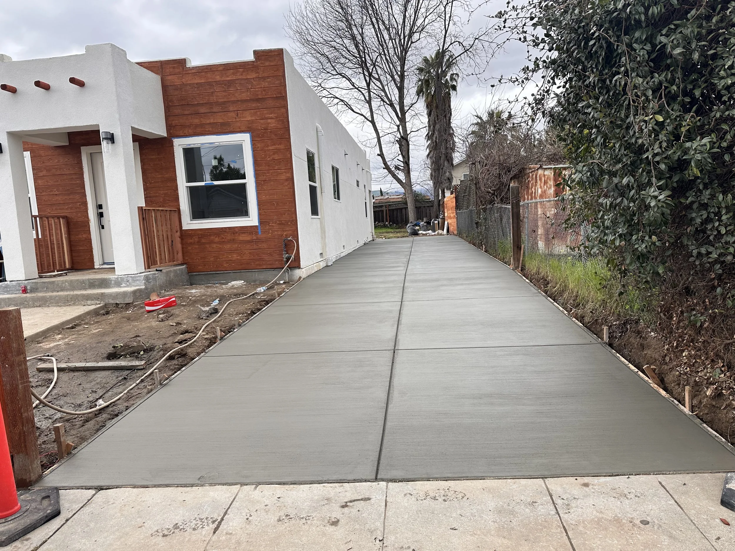 Newly poured concrete driveway next to a modern house with white walls and wooden accents, with unfinished landscaping and construction materials nearby.