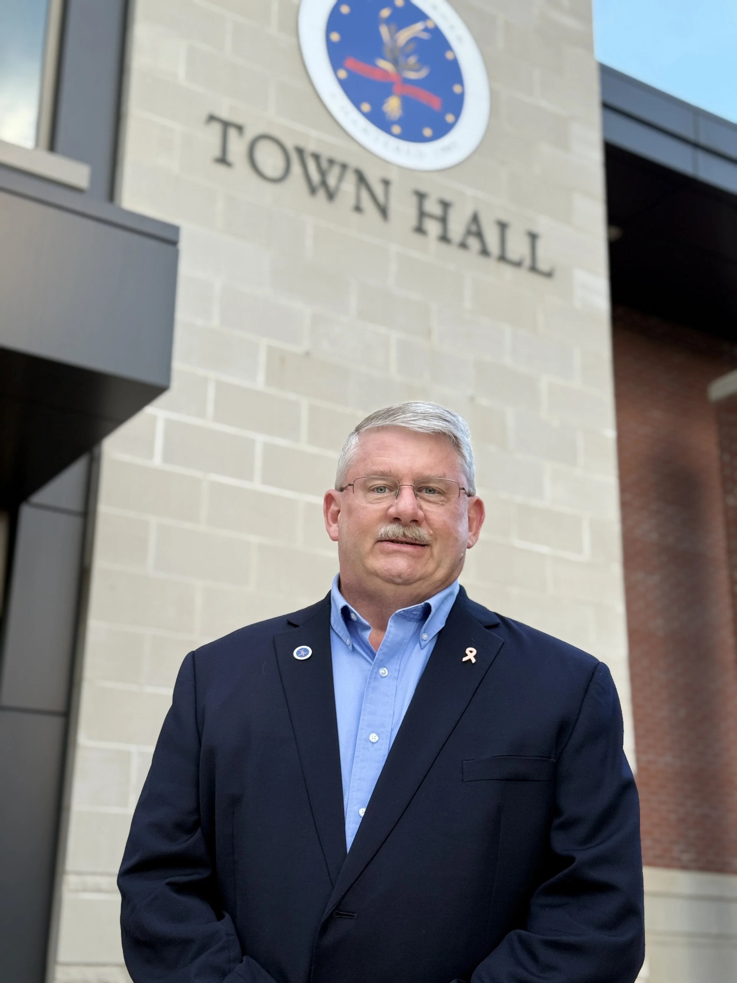 A man in a blue suit jacket and light blue shirt standing in front of Town Hall building with a clock and a sign.