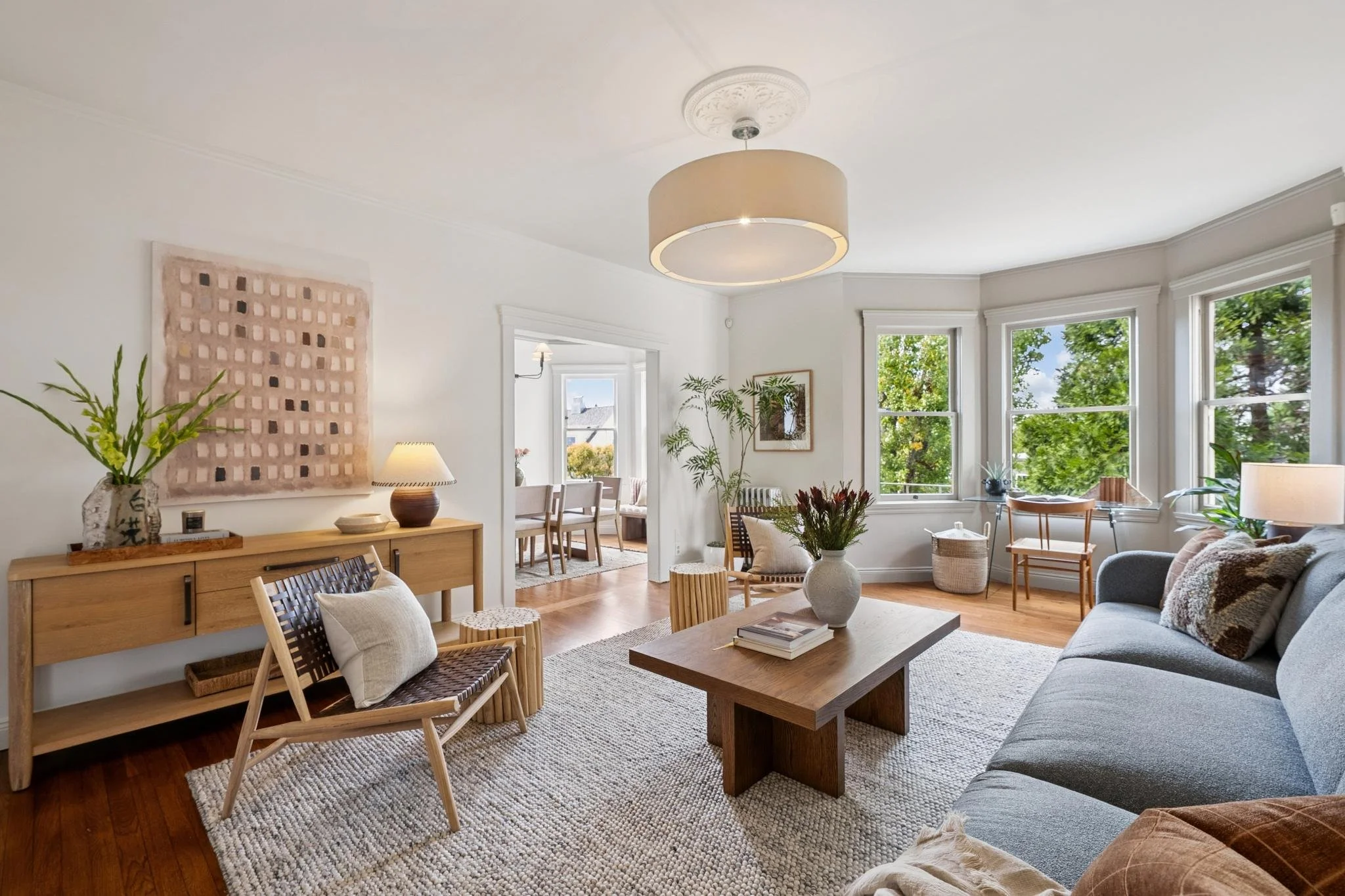 Bright living room with large windows, beige and gray furniture, wooden coffee table, potted plants, wall art, and a round ceiling light