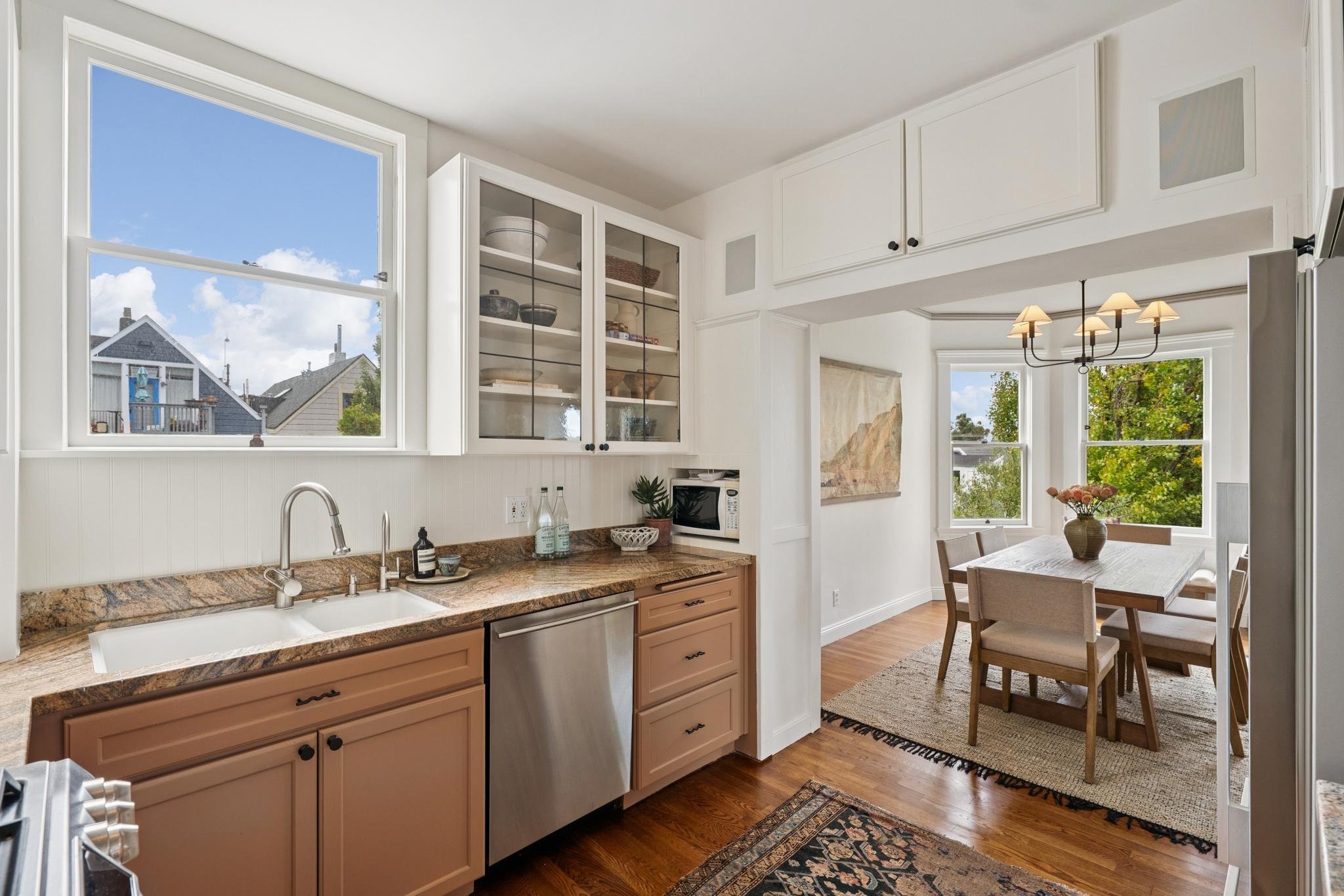 View of a kitchen with beige cabinets, granite countertops, and a white sink. There are large windows letting in natural light, and a dining area with a wooden table, chairs, and a vase with flowers. The ceiling has a chandelier with multiple lamps.