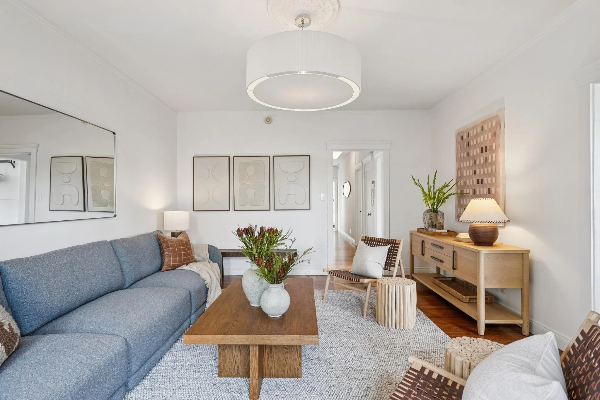 Living room with white walls, wooden flooring, a blue sofa with pillows, wall art, a wooden coffee table with a plant, a side table with a lamp, and additional decorative furniture and plants.