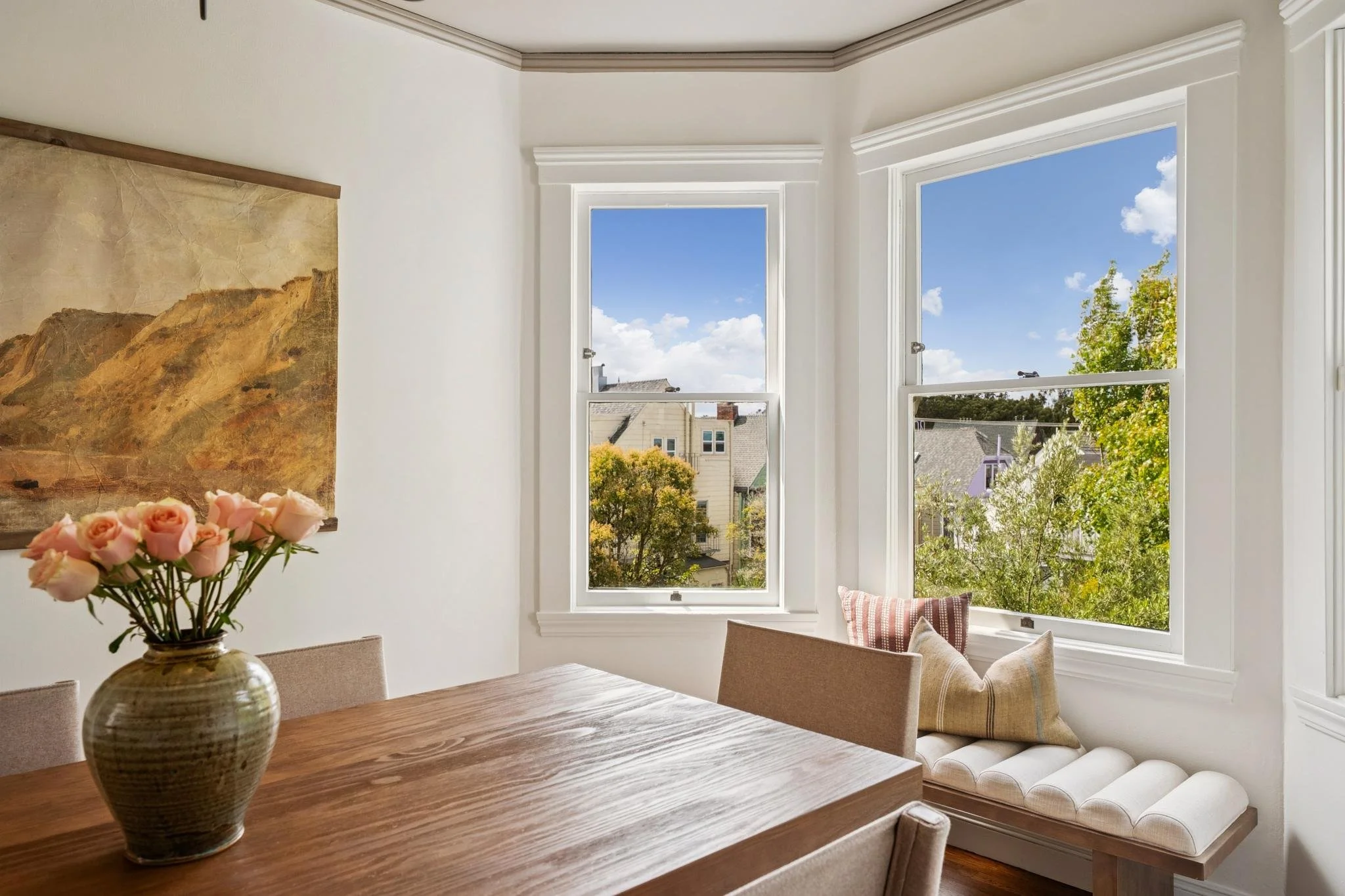 A cozy dining nook with a wooden table, a beige cushioned bench, and a vase of pink roses, with three large windows showcasing a view of houses and green trees outside on a bright, sunny day.