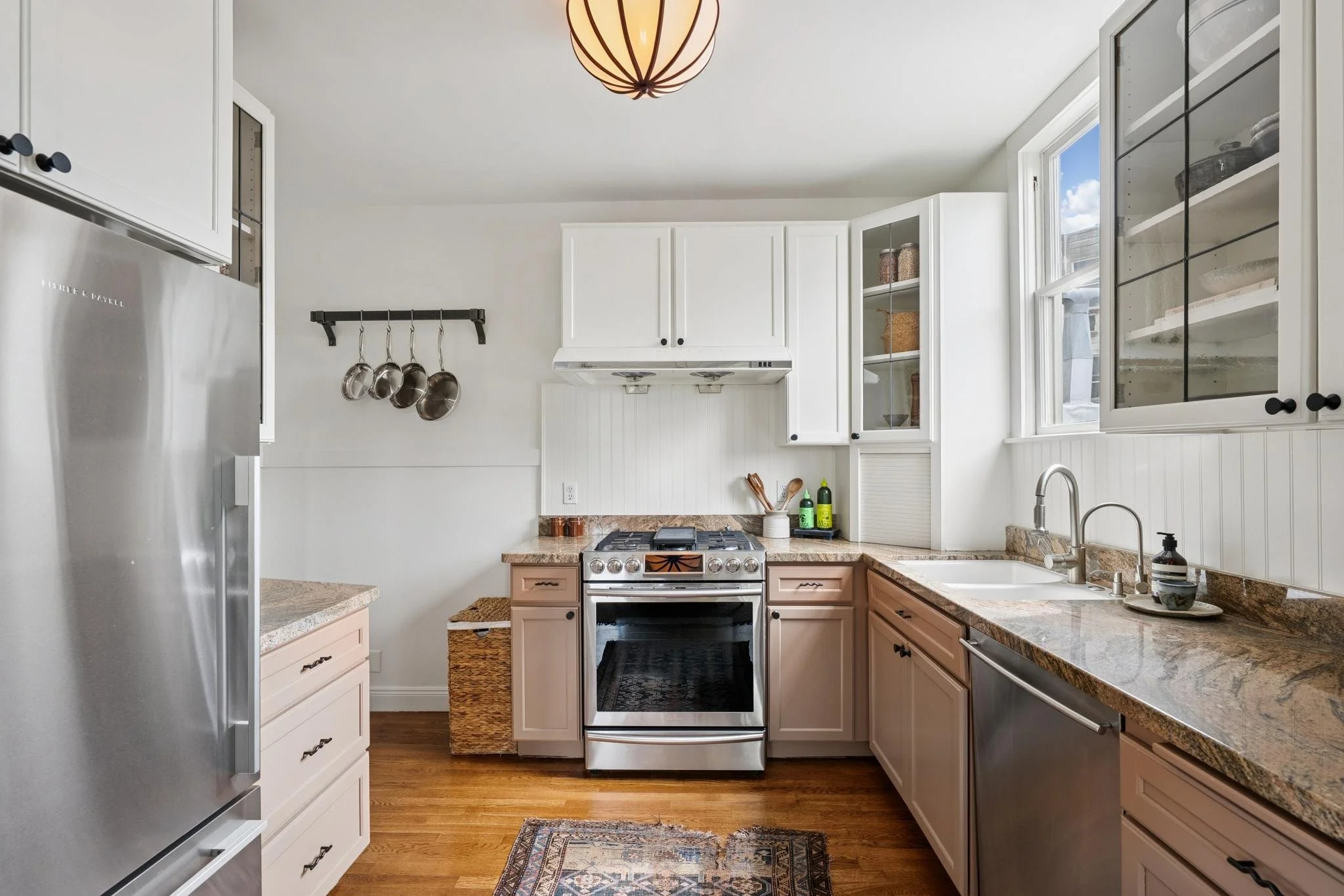 A bright kitchen with white cabinets, a stainless steel refrigerator, a beige granite countertop, and a window letting in natural light. There is a gas stove in the center, and hanging pots on the wall. A small rug is on the wood floor, and various k