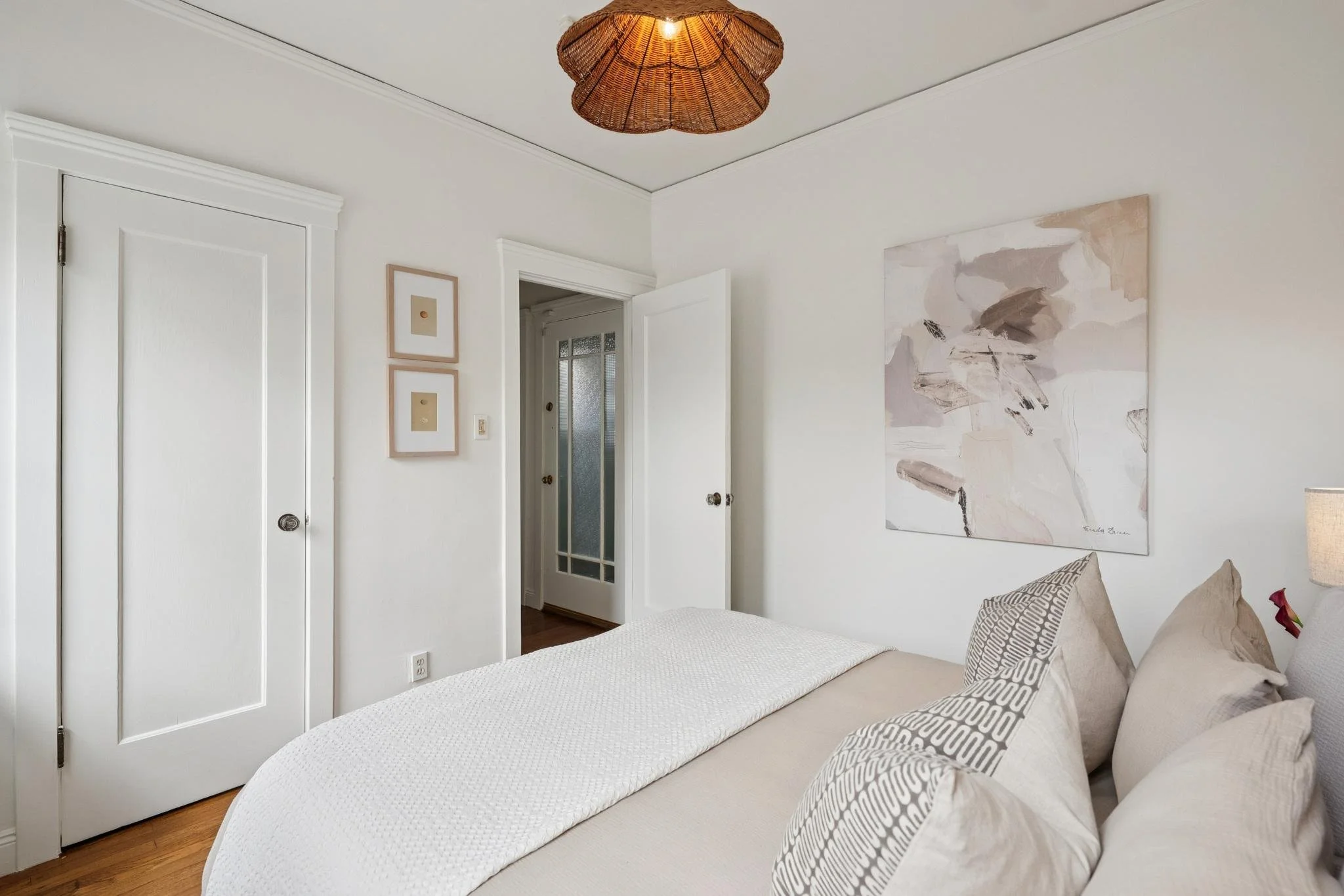 A bedroom with white walls, a white bedspread, and multiple pillows with patterned pillowcases. There are two small framed pictures on the wall, an abstract painting, and a wicker ceiling light.