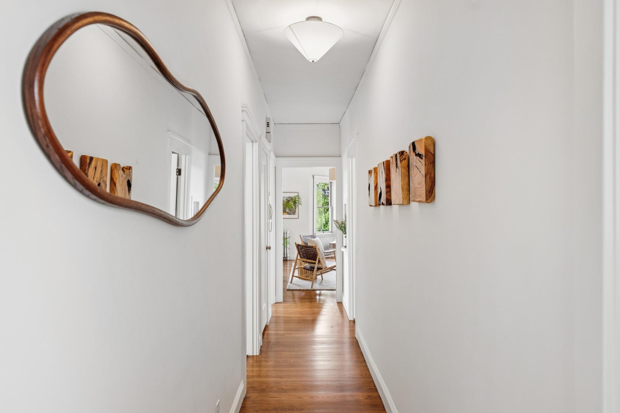 A hallway in a home with white walls and hardwood flooring, decorated with wooden wall art and a curved mirror, leading to a room with chairs and a window.