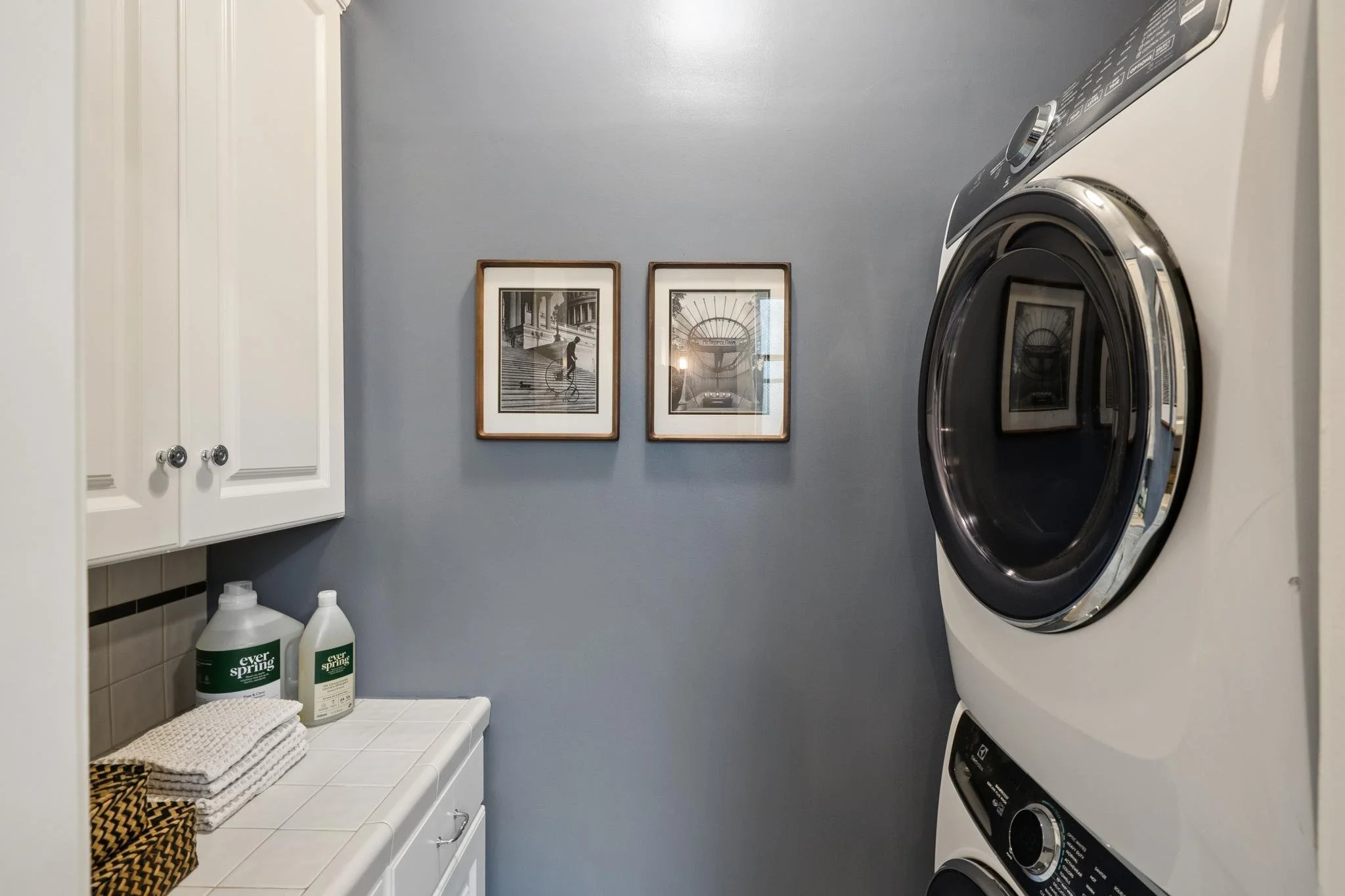 A laundry room with grey walls and white cabinets, containing a stacked washer and dryer, framed black and white photos on the wall, two bottles of laundry detergent, and a neatly folded towel on a white countertop.