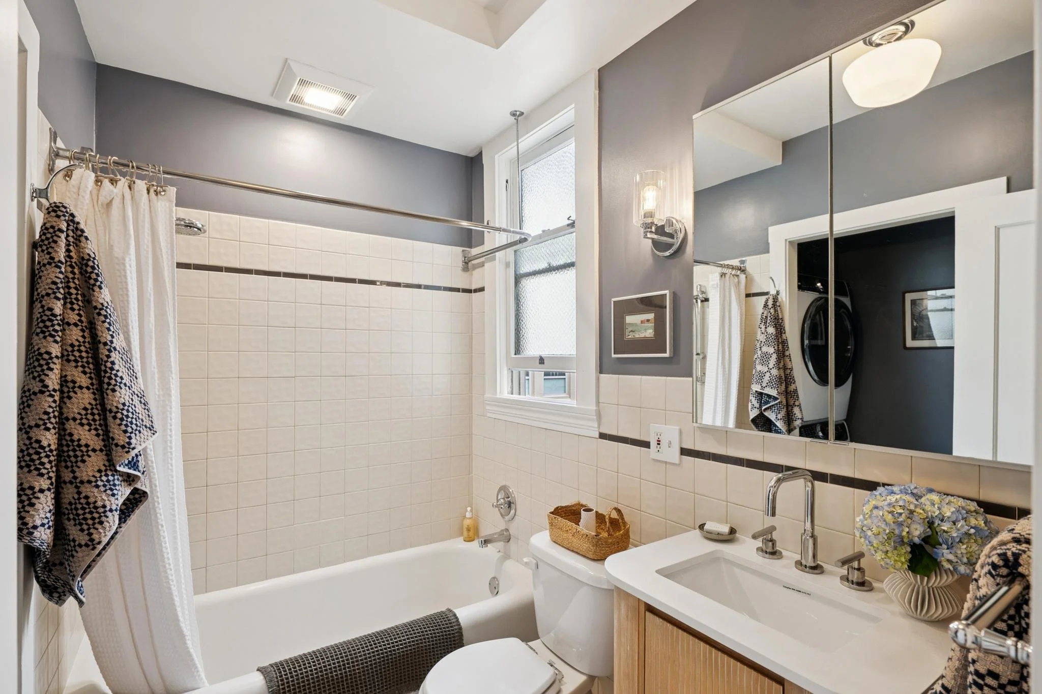 A modern bathroom features a bathtub with a tiled wall, a window with frosted glass, a white sink with a faucet, a large mirror, floral decor, a basket with toiletries, and a window with natural light.