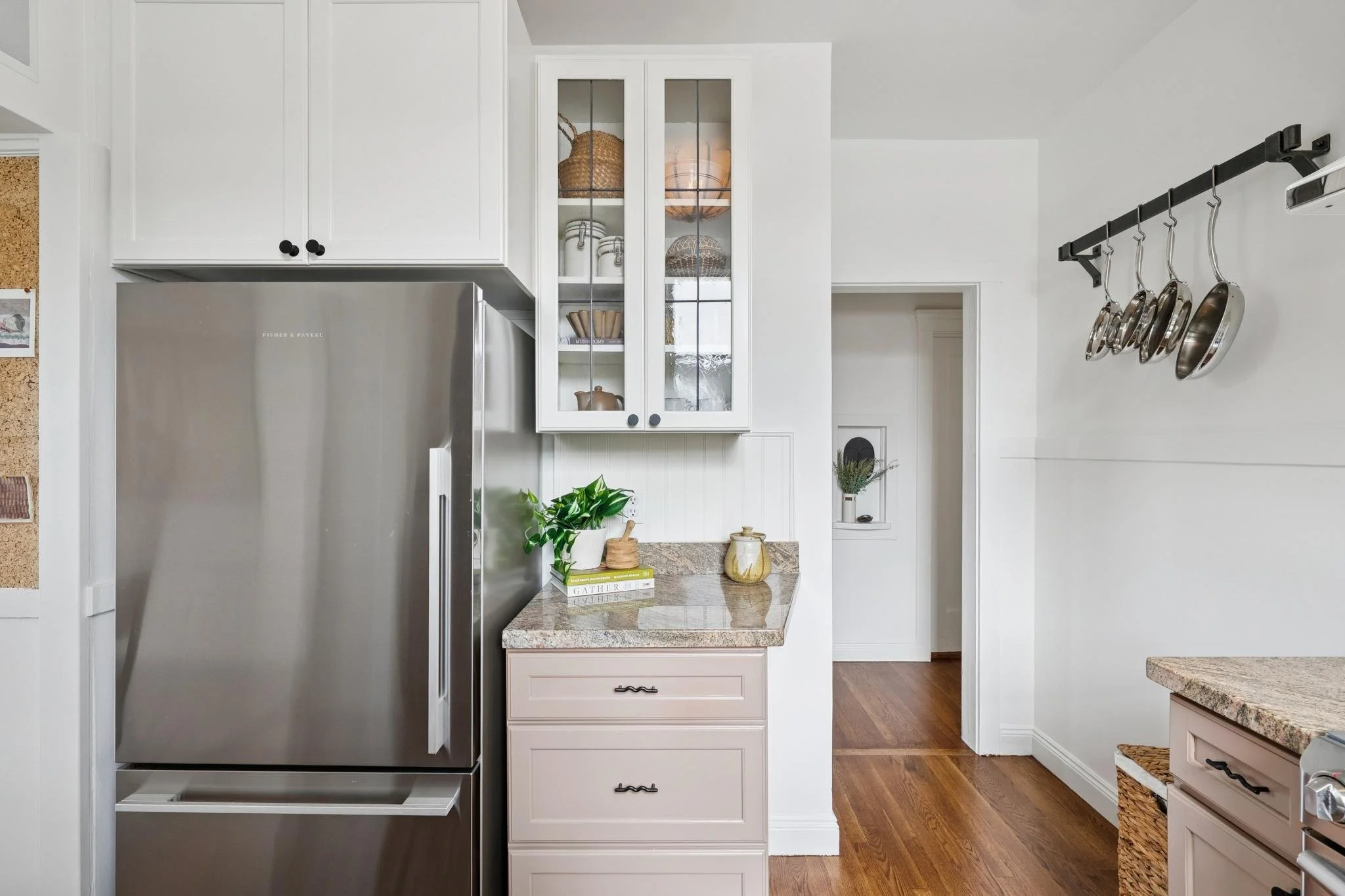 A modern kitchen with white cabinetry, a stainless steel refrigerator, and a granite countertop. There is a glass-front cabinet displaying baskets and dishes, and a black pot rack with hanging pots and pans. Wooden flooring is visible.