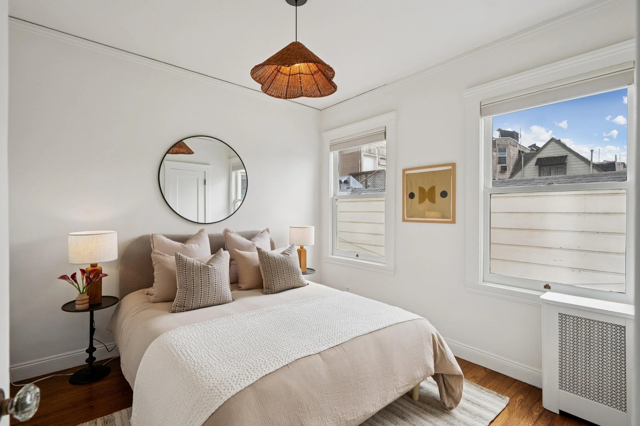 A cozy bedroom with a beige bed, multiple pillows, and a white blanket, flanked by two side tables with lamps, and a large mirror above the headboard. There are two windows with white frames showing neighboring houses and a blue sky outside, and simp