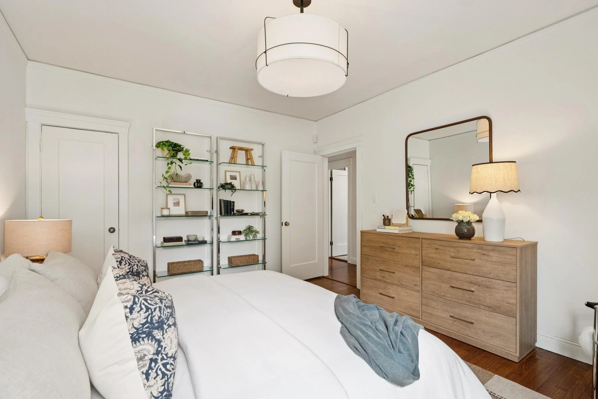 A bedroom with a white bed, a wooden dresser, a mirror, a lamp, and a shelving unit with decorative items in a bright, minimalist style.