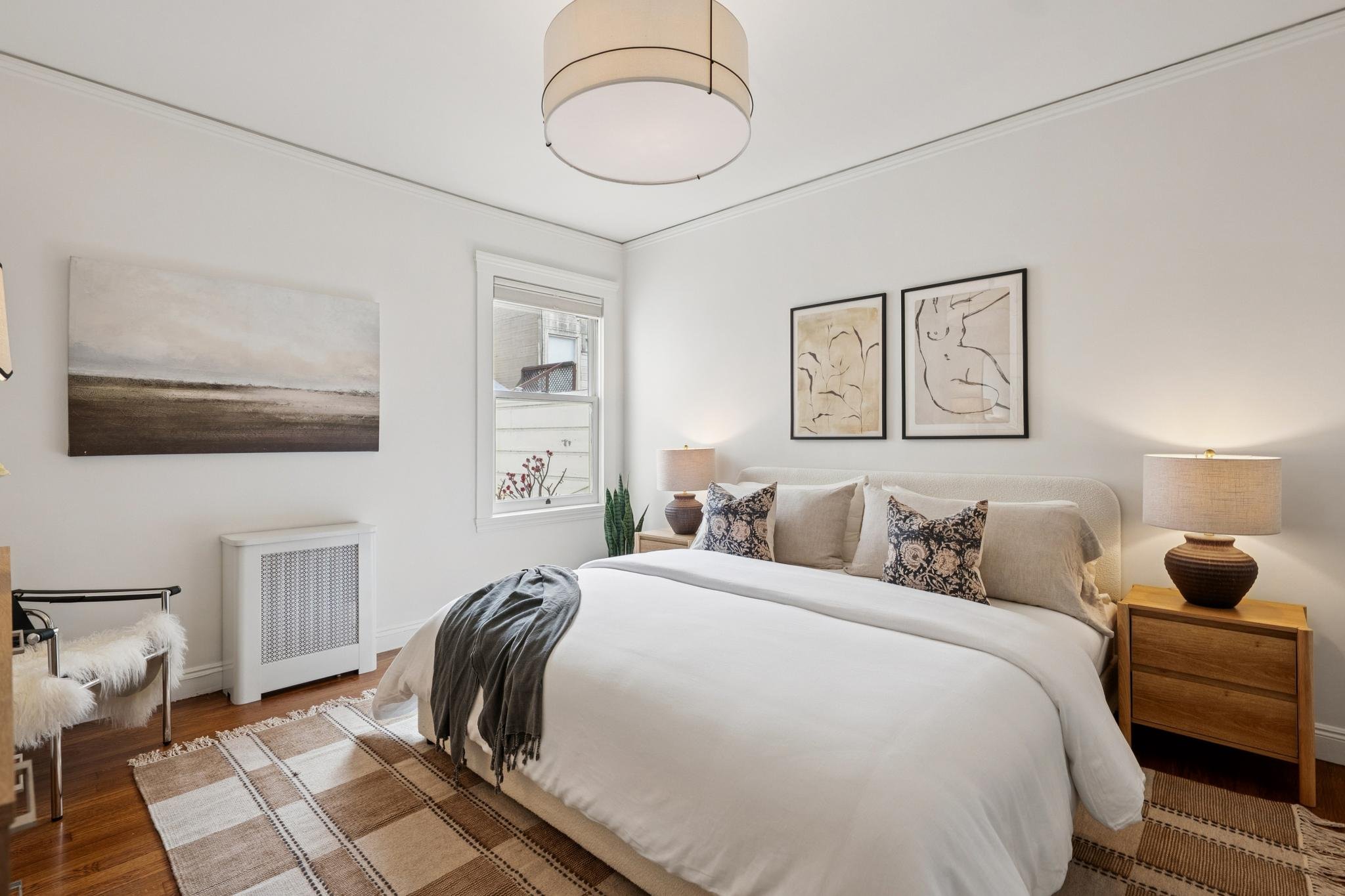 A cozy bedroom with white walls, a large bed with white bedding and patterned pillows, wooden nightstand with a lamp, artwork on the walls, a window, and a small chair with a furry cushion.