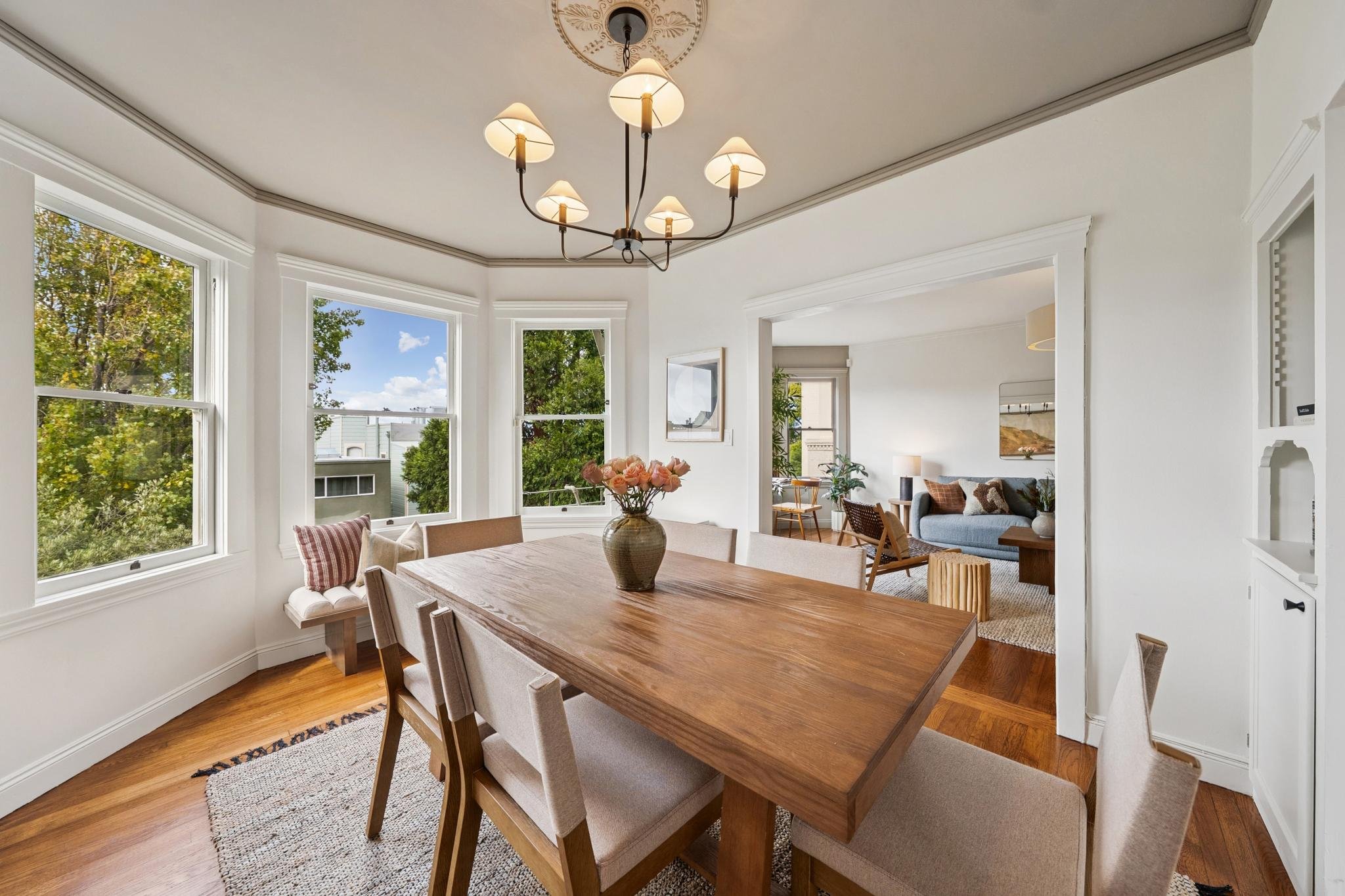 A bright dining area with a wooden table and beige chairs, a floral centerpiece, large windows showing green trees and a blue sky, and a view into the living room with a gray couch and art on the wall.