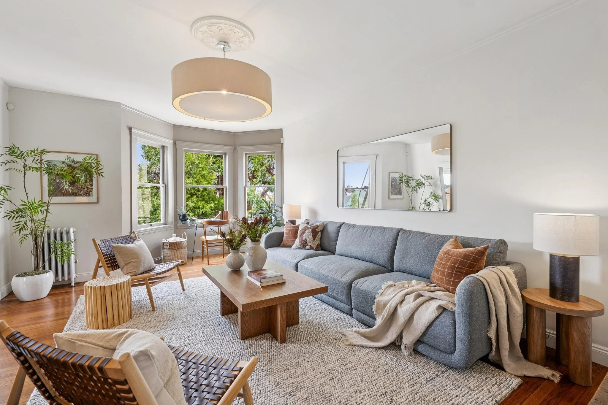 Living room with a large gray sectional sofa, wooden coffee table, multiple patterned pillows, plants, wall art, and large windows with greenery outside.