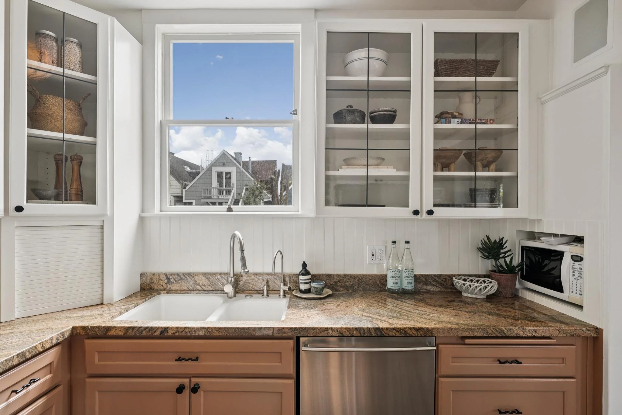 View of a kitchen with white cabinets, a granite countertop, a window showing neighboring houses and a blue sky, a stainless steel sink and faucet, decorative bowls and bottles, a small microwave, and a plant.