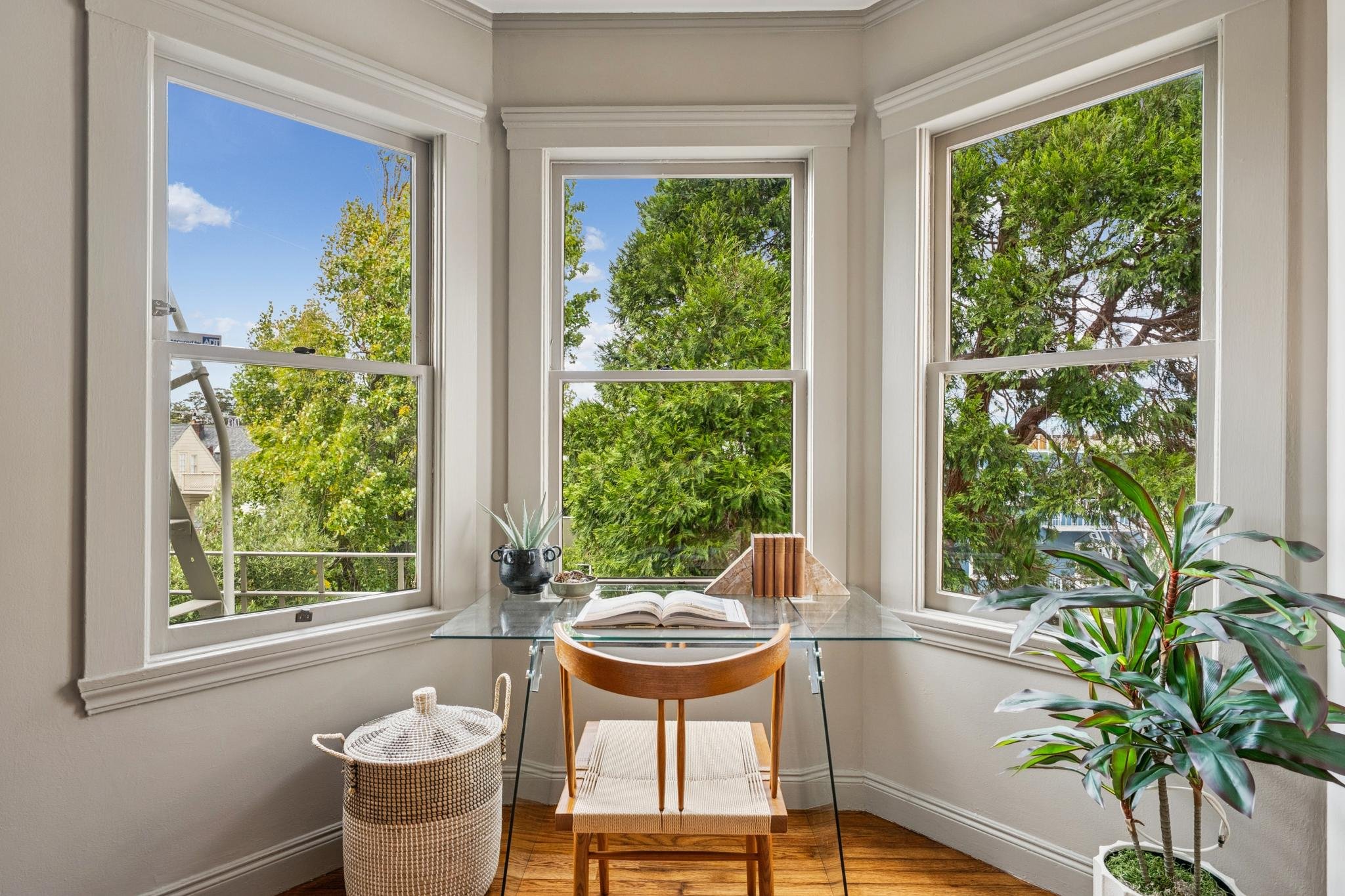 A cozy reading nook with a glass-topped desk, surrounded by large bay windows with white trim, overlooking trees and a blue sky. Decor includes potted plants, a closed book, and a stool with a woven basket nearby.