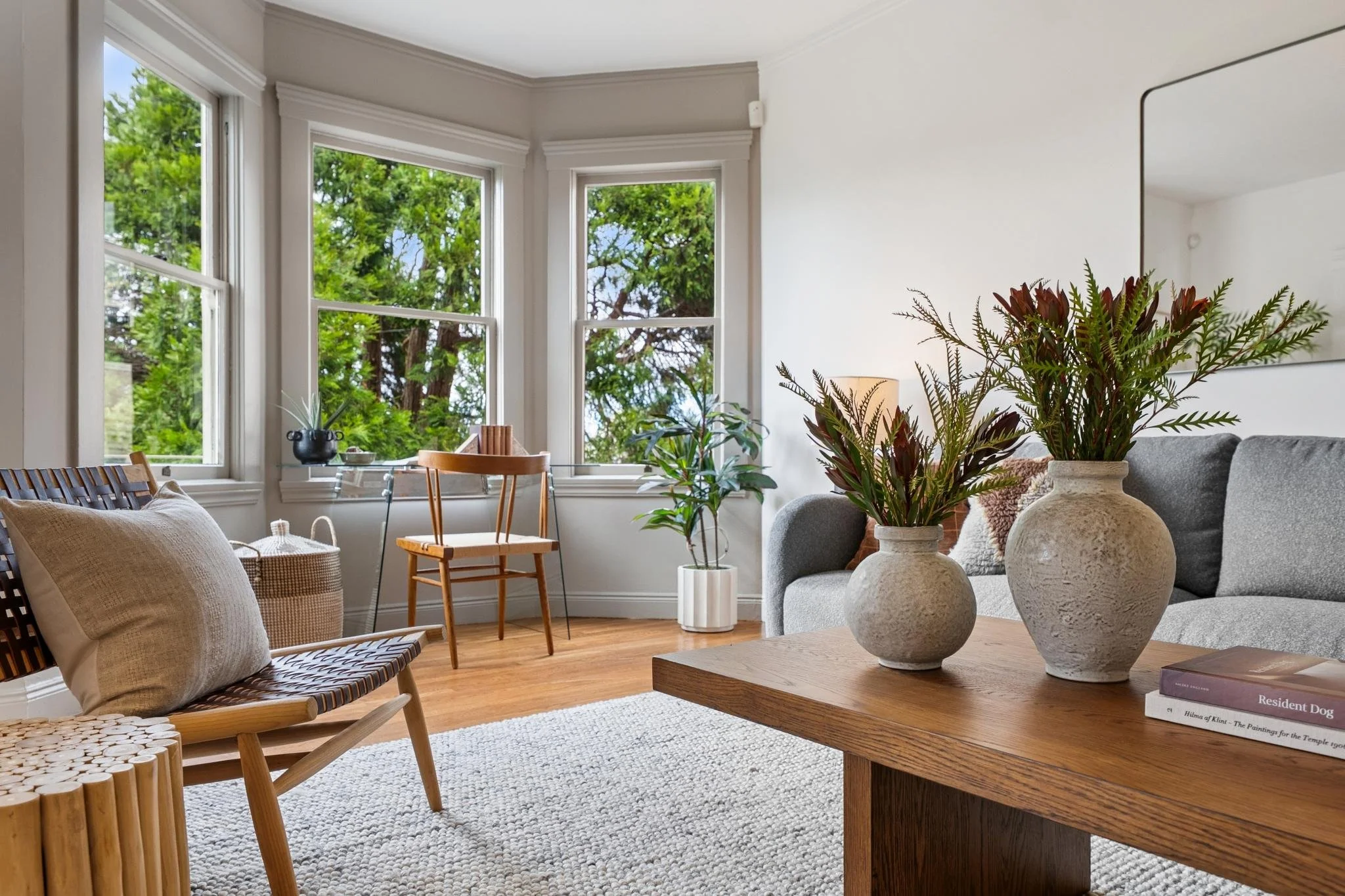 Living room with large windows, potted plants, a beige armchair, a wooden bench, a gray sofa, and decorative vases on a wooden table.