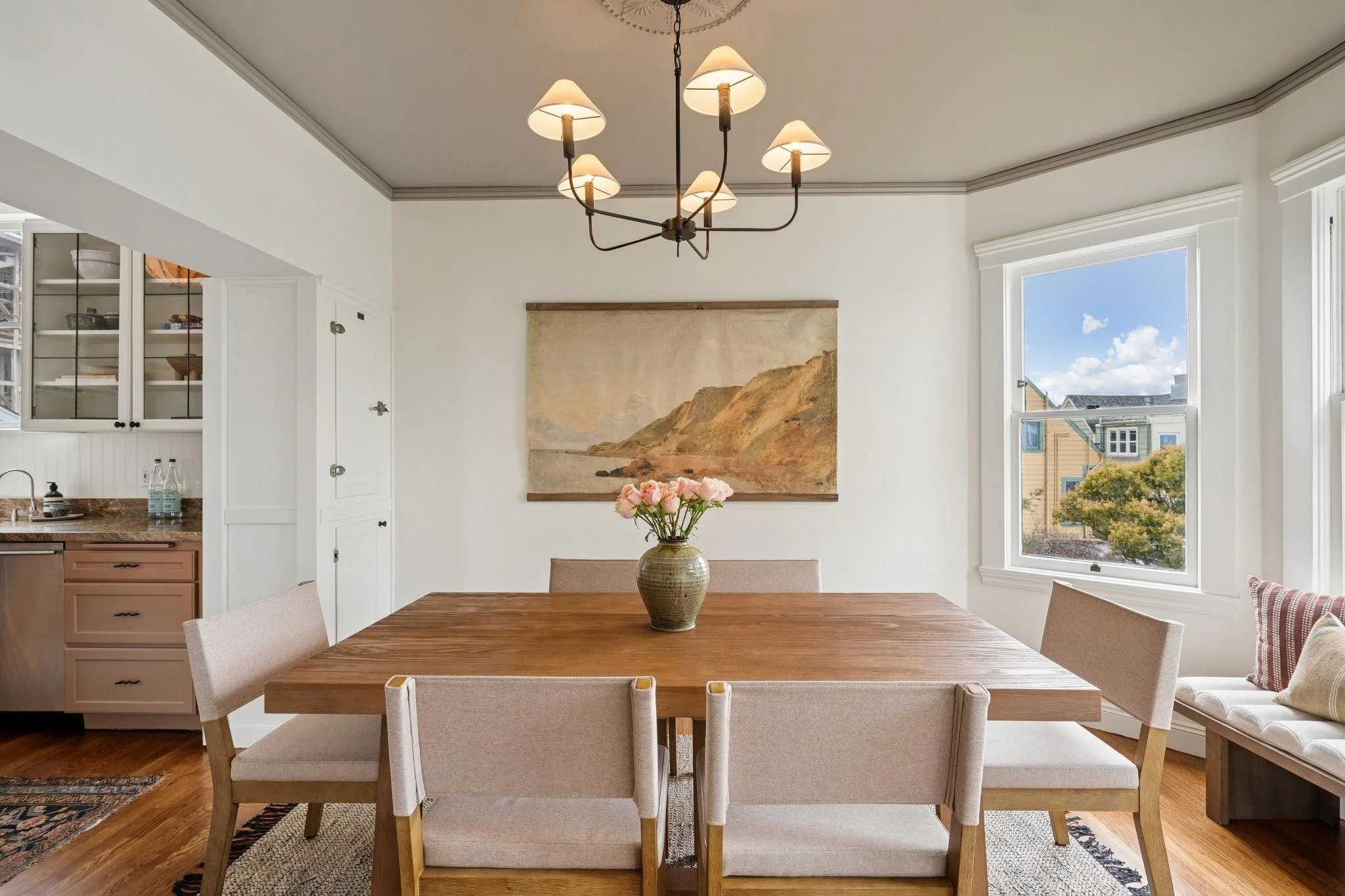 A dining room with a wooden table, beige upholstered chairs, a vase of pink flowers, large window with a view of houses and trees, artwork on the wall, and a chandelier with seven lamps.