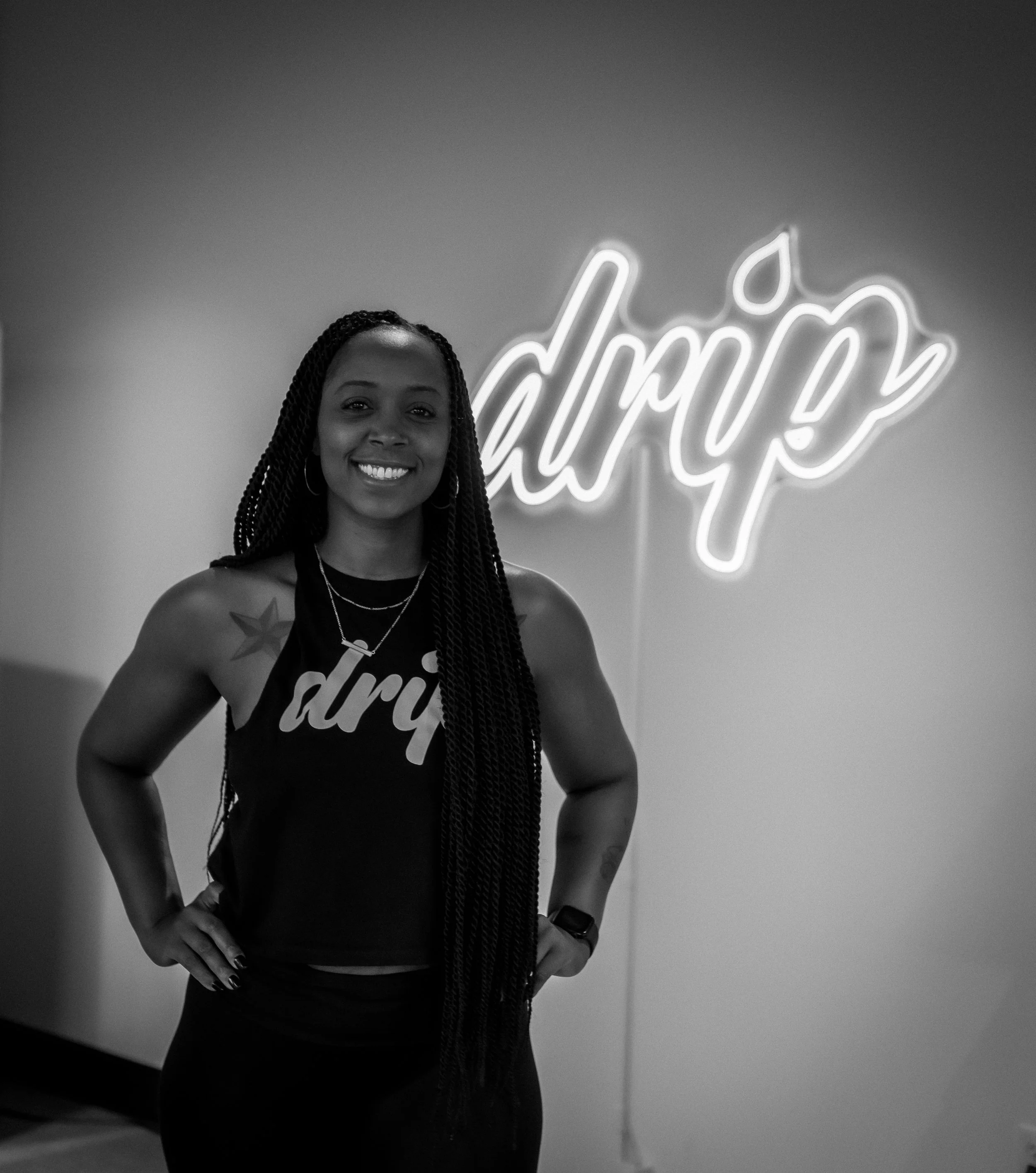 A smiling woman with long braids wearing a sleeveless shirt with the word 'driq' in front of a neon sign that says 'driq' with a leaf design.