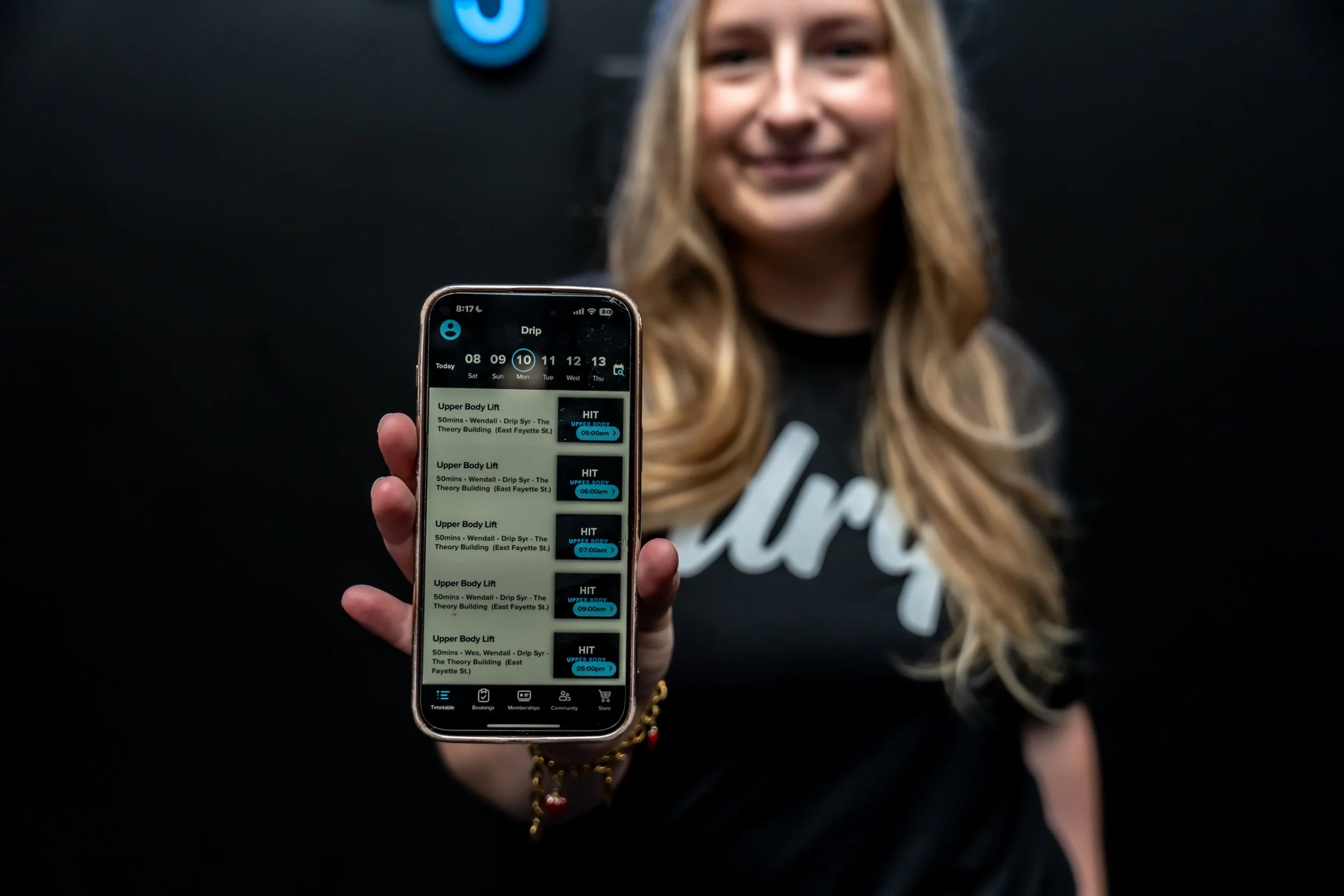 A woman holding a smartphone displaying a fitness class schedule app, with her face blurred and a black background.