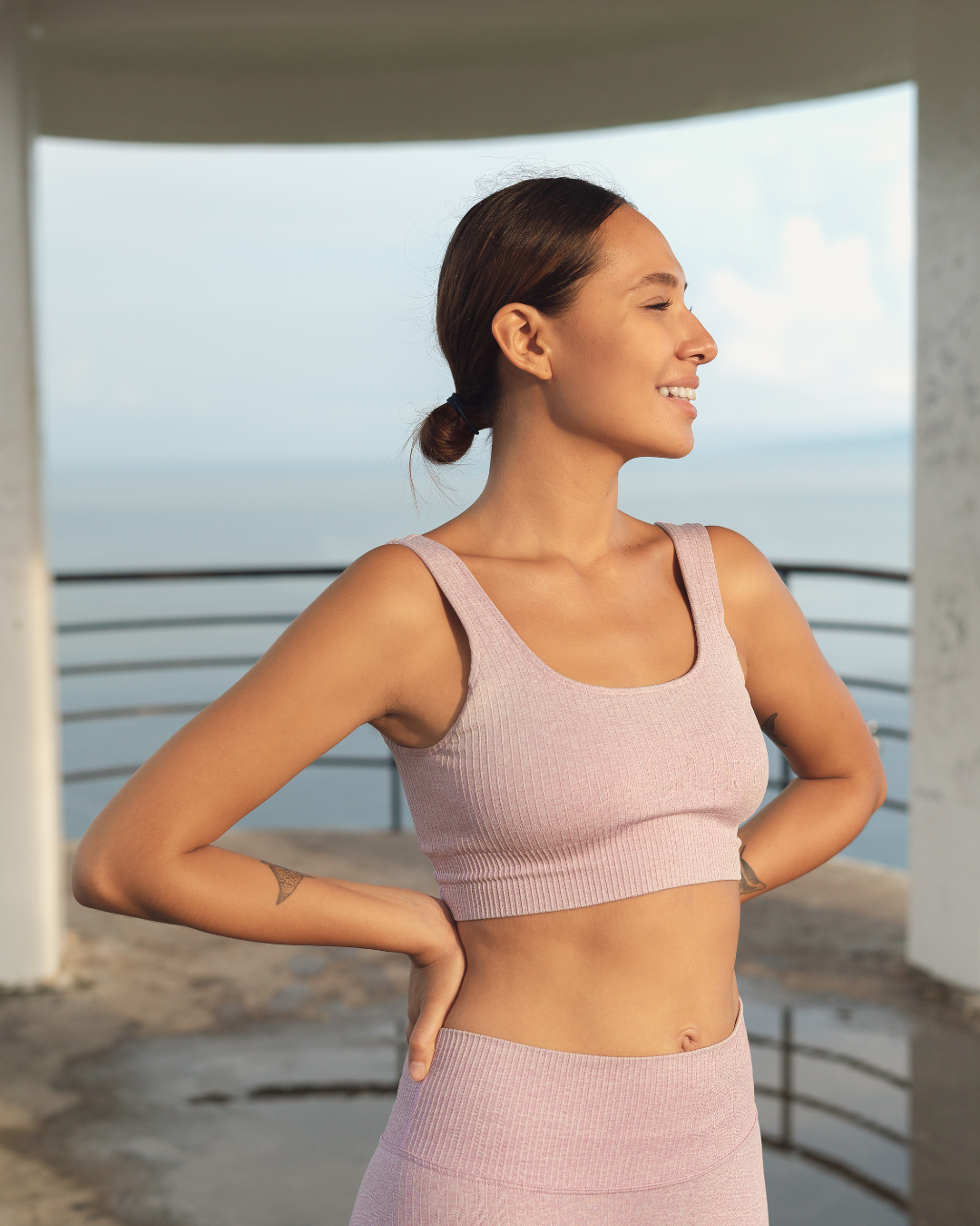 A woman with brown hair tied back, wearing a pink sports bra and matching high-waisted leggings, standing outdoors on a pier or balcony, smiling and looking to the side with ocean in the background.
