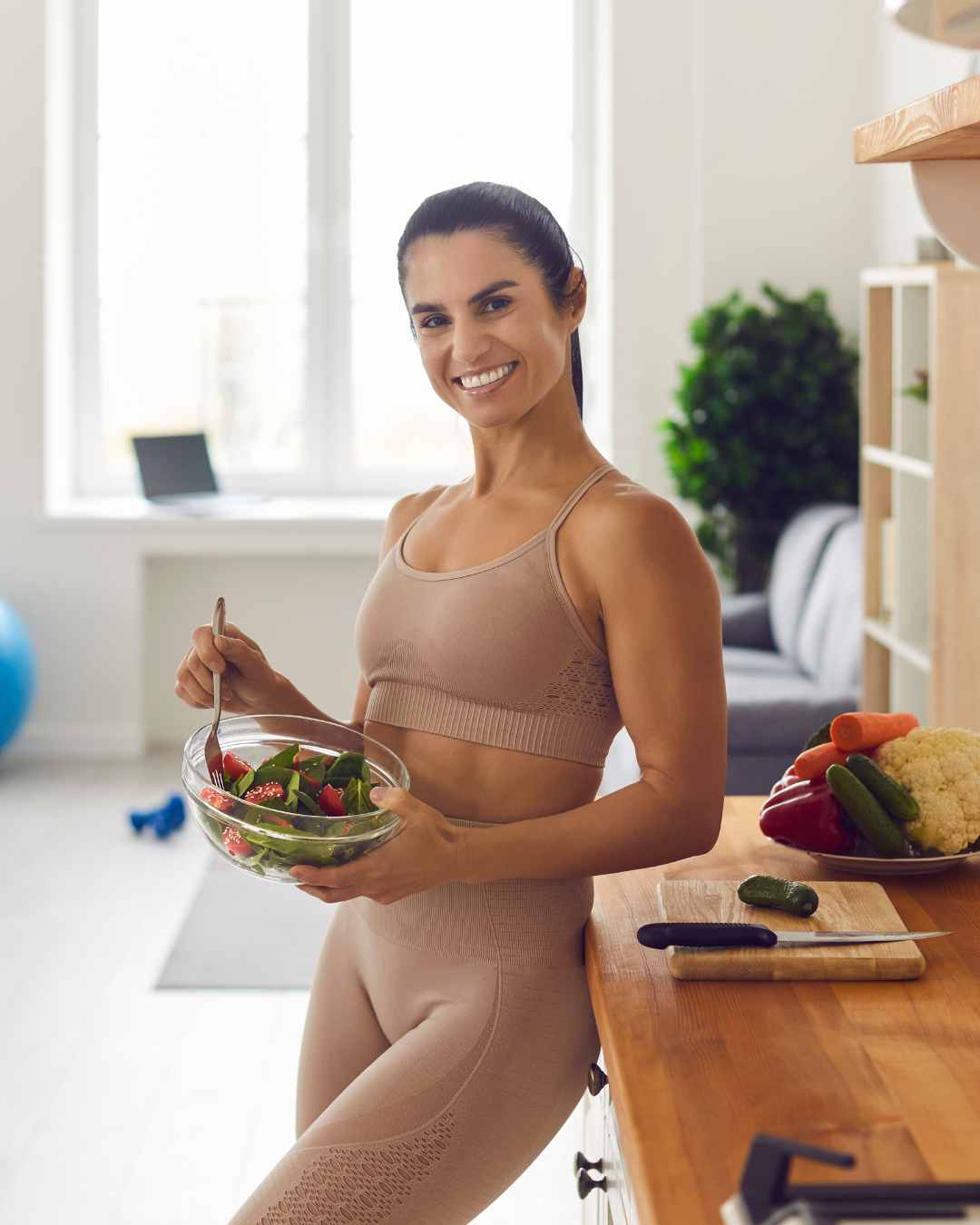 Smiling woman in beige workout clothes preparing a salad in a bright kitchen with fresh vegetables on the counter.