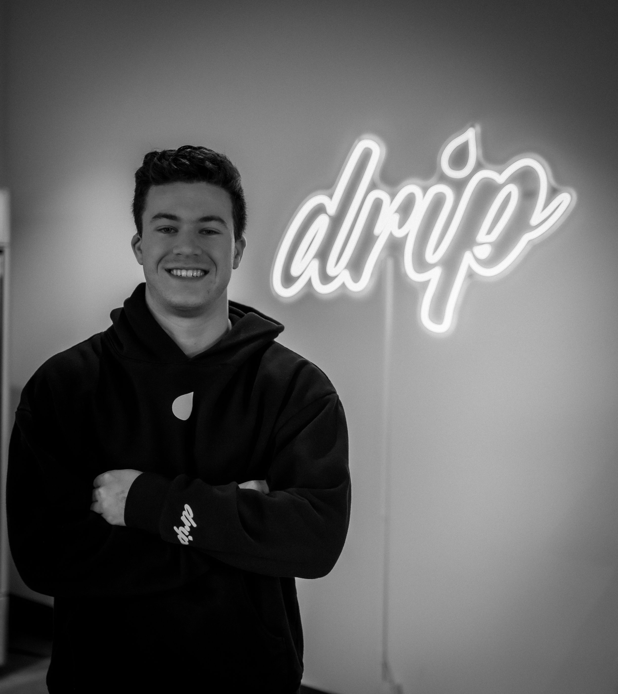 A young man with dark hair smiling and crossing his arms, standing in front of a neon sign that says 'dri' with a water droplet icon, in a black and white photo.