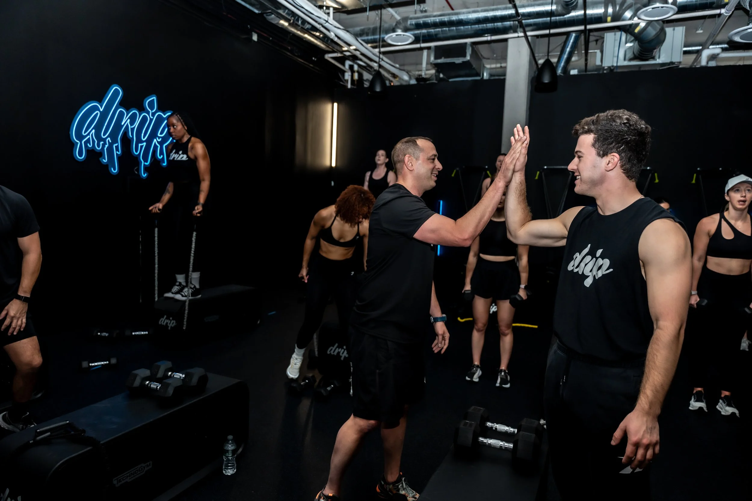Two men high-fiving during a workout class in a gym with women exercising in the background.