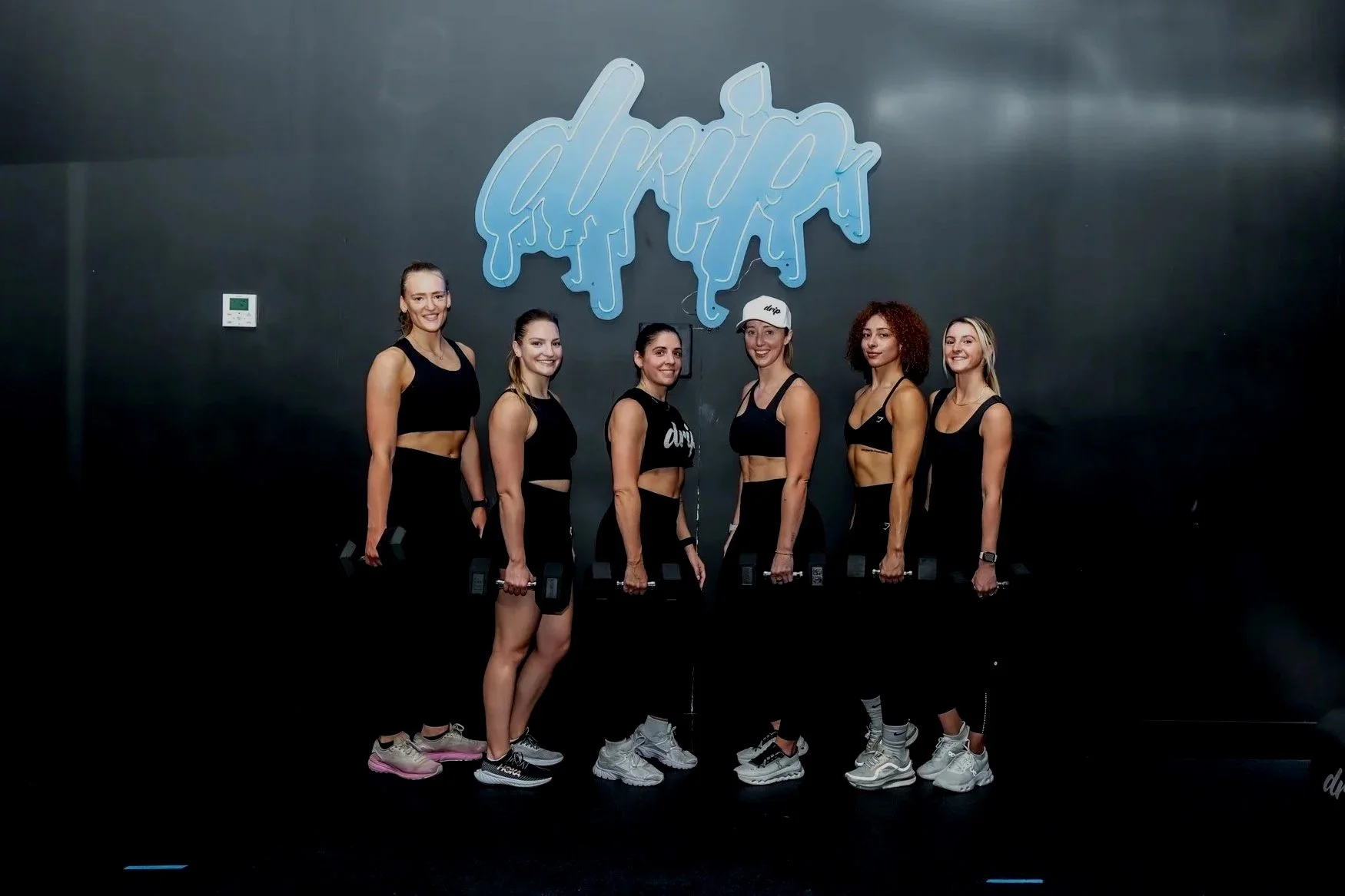 Six women standing in a line, holding dumbbells during a workout session at a gym with a dark wall and a blue logo that says 'drip' in the background.