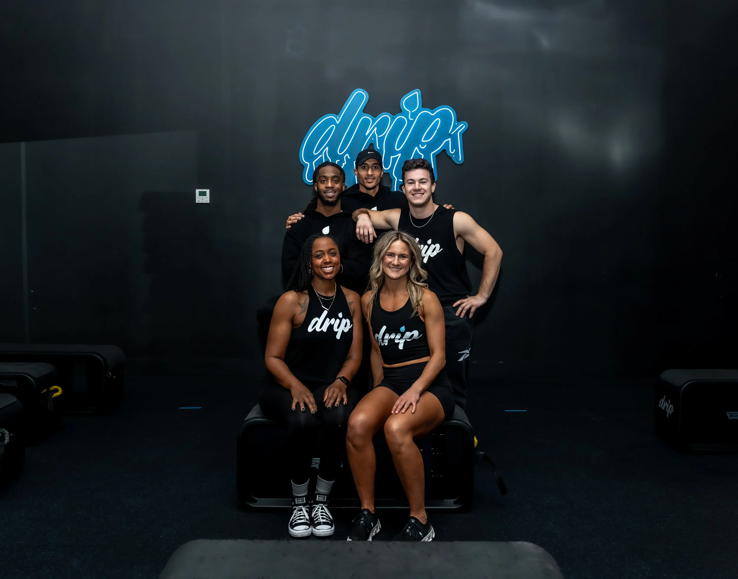 Group of six diverse people posing together in a fitness studio with a neon sign that says 'drink' on a black wall. Two women are sitting on black workout benches, three men are standing behind their chairs, and one man is standing in the middle behind the women. They are all smiling and wearing workout clothes with the word 'drip' on some of their tops.