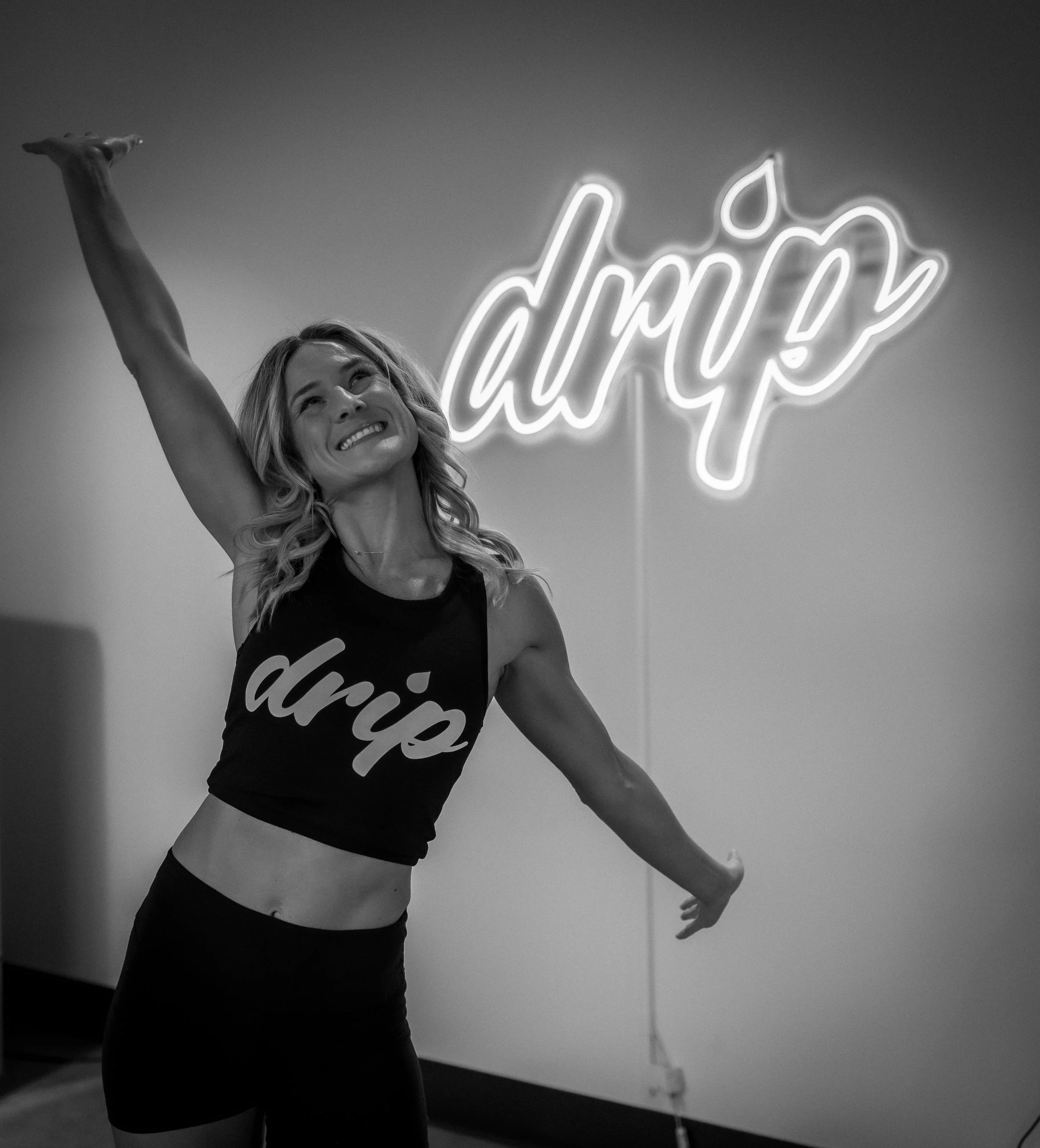 A woman smiling and stretching her arms in front of a neon sign that says 'dripe' with a leaf design.