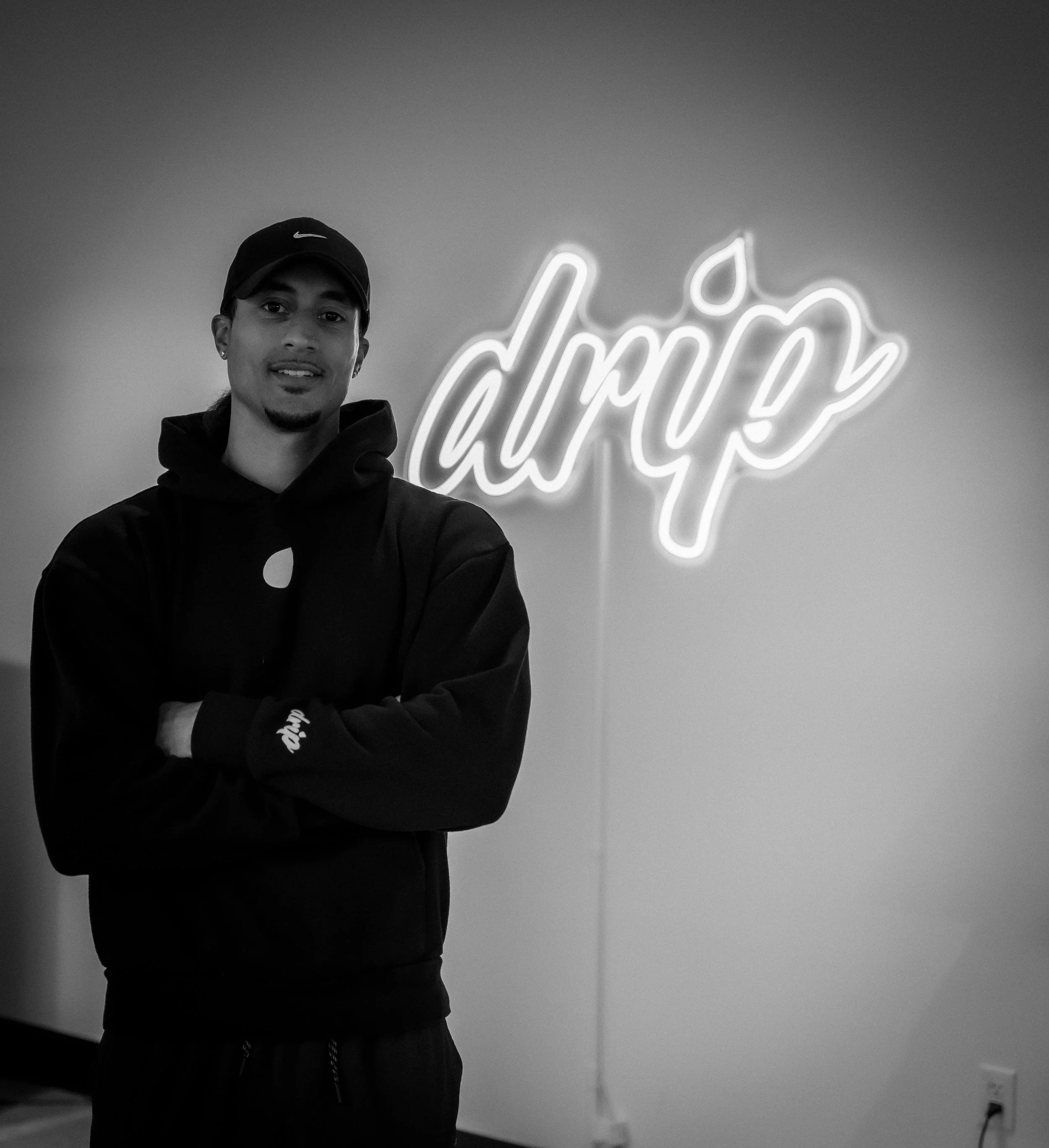 A young man in a black hoodie and cap standing with arms crossed in front of a wall with a neon sign that says 'dayp' in cursive.