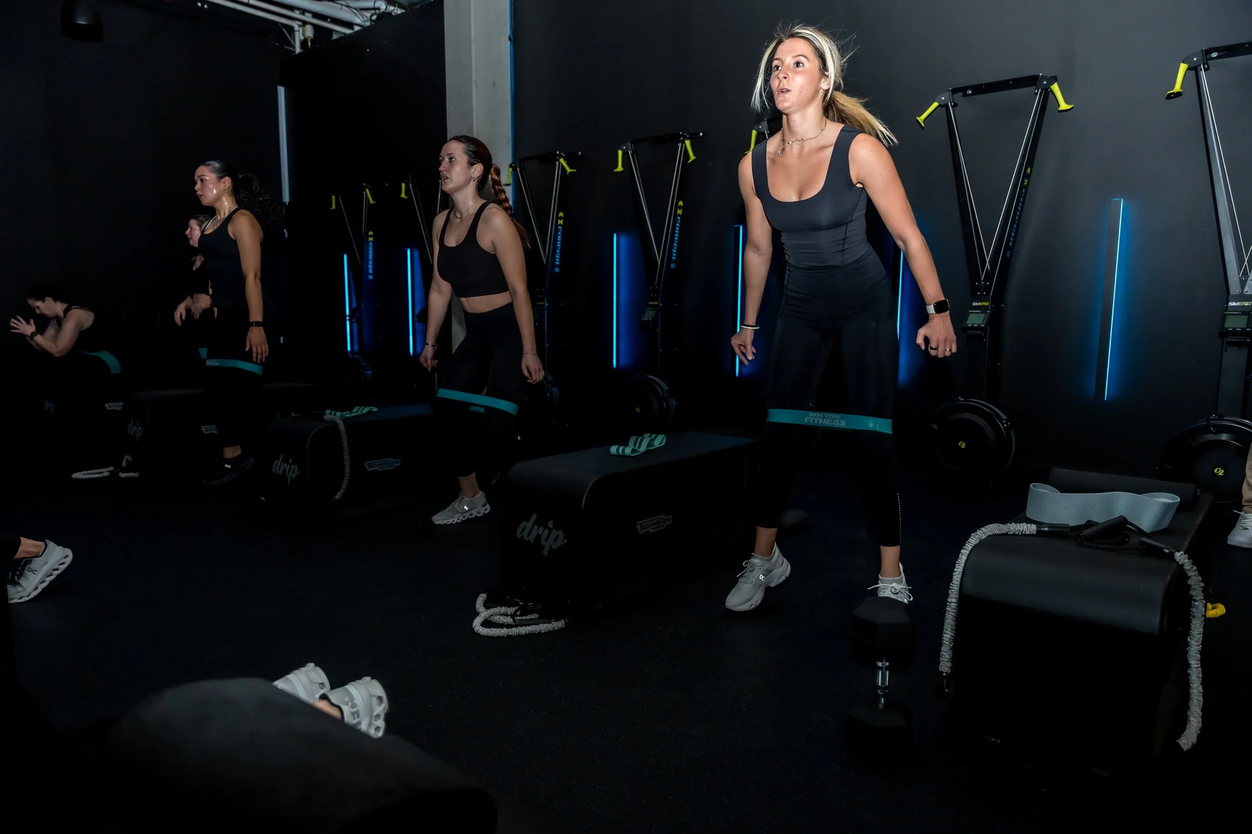 Women participating in a group fitness class in a dark gym with black walls, doing strength exercises with weighted bags, resistance bands, and bodyweight movements.